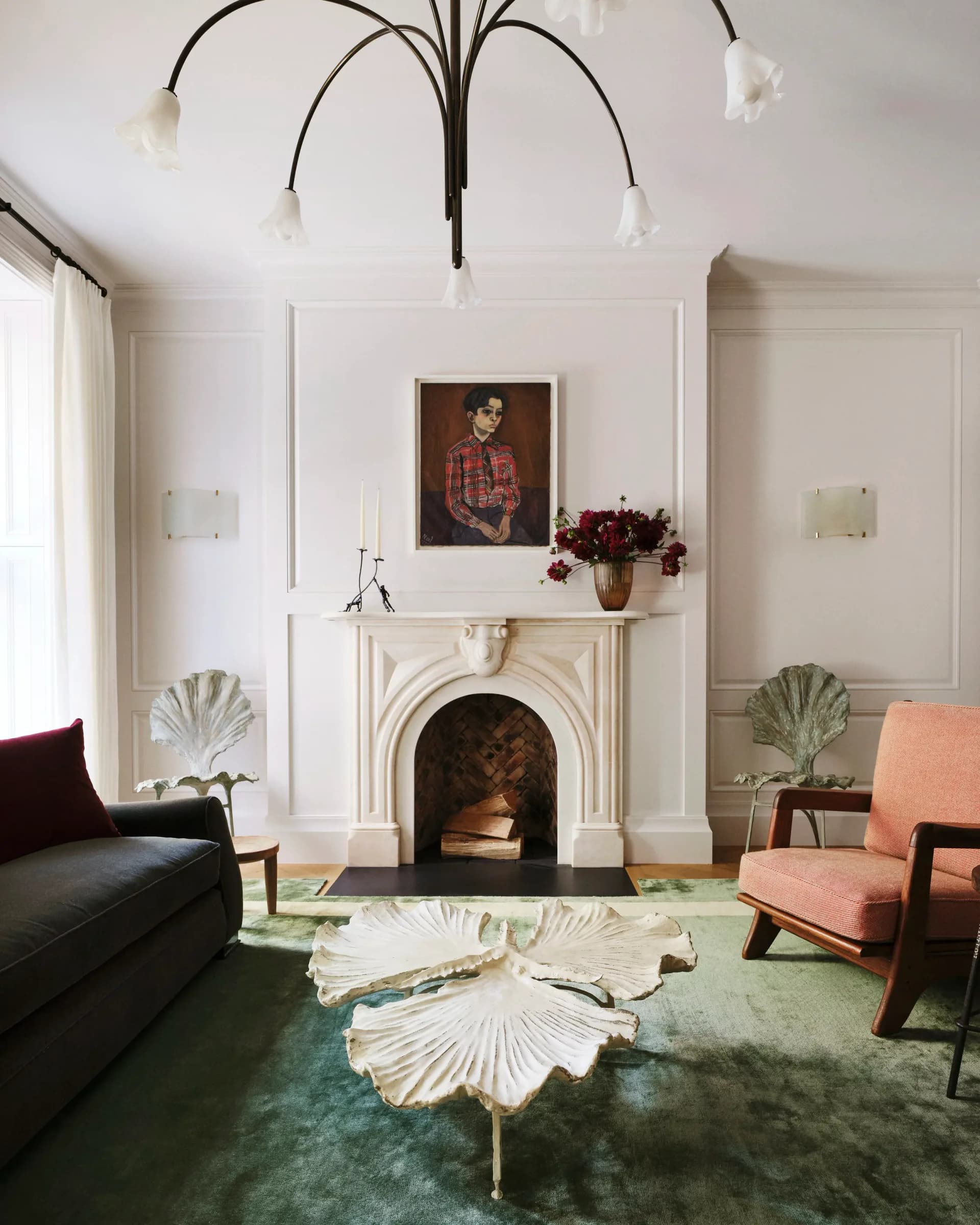 Elegant living room with white fireplace mantel, black chandelier, green patterned rug, coral chair, and shell-shaped coffee table by Ashe Leandro.