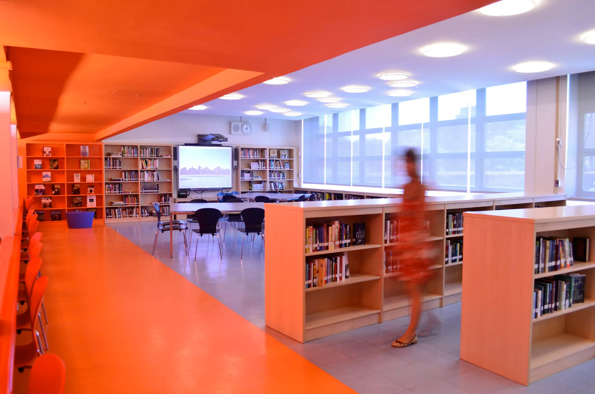 Vibrant library interior with bold orange flooring, ceiling, and built-in shelving, blue accent lighting, and large windows, designed by Atelier Pagnamenta Torriani.