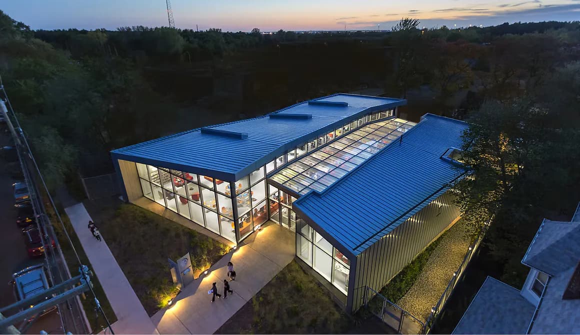 Modern industrial building with bright blue corrugated metal roof and glass facade, illuminated at dusk surrounded by trees, designed by Atelier Pagnamenta Torriani.