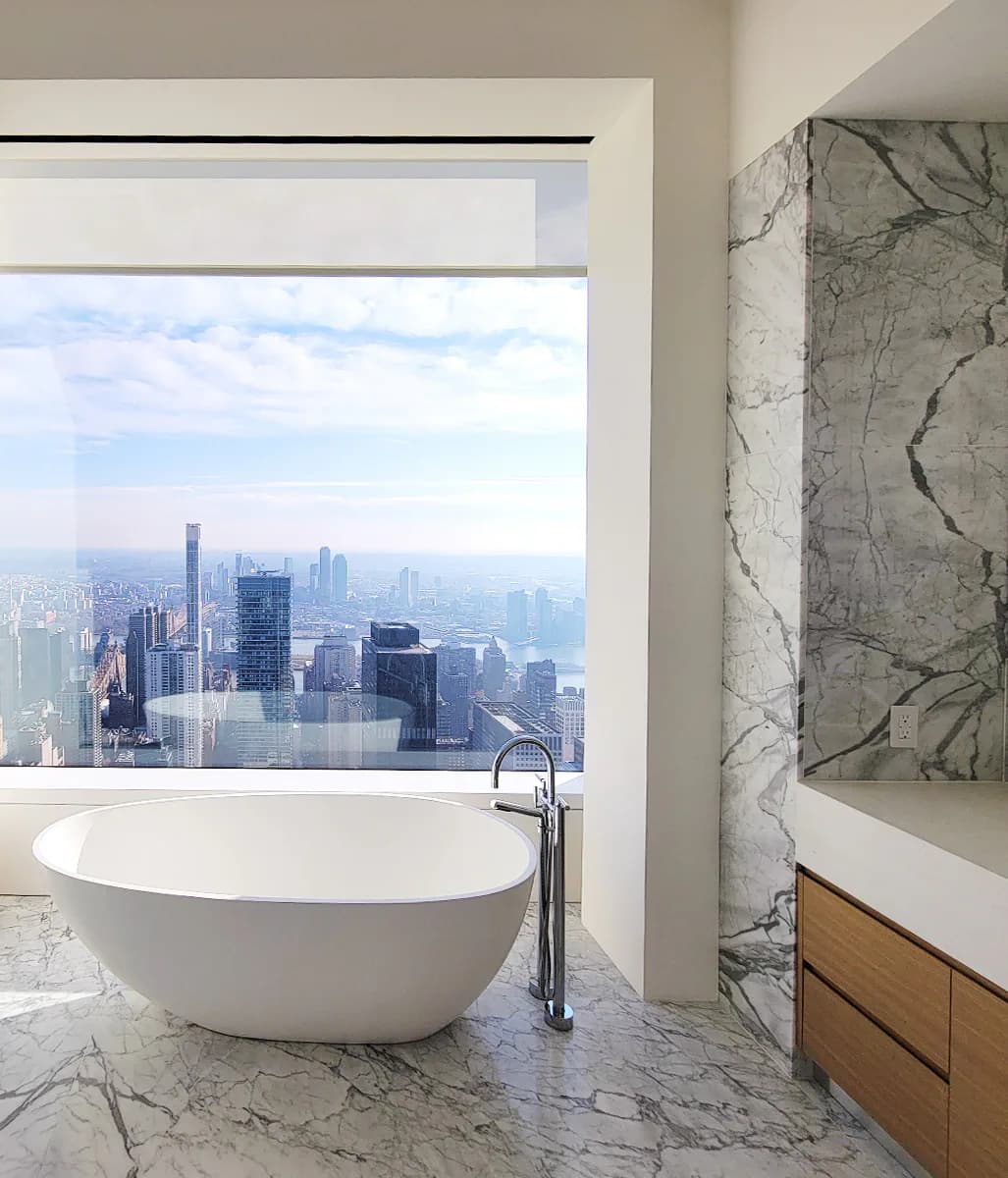 Luxurious marble bathroom with freestanding white oval tub overlooking Manhattan skyline through floor-to-ceiling windows, designed by Atelier Premiere.