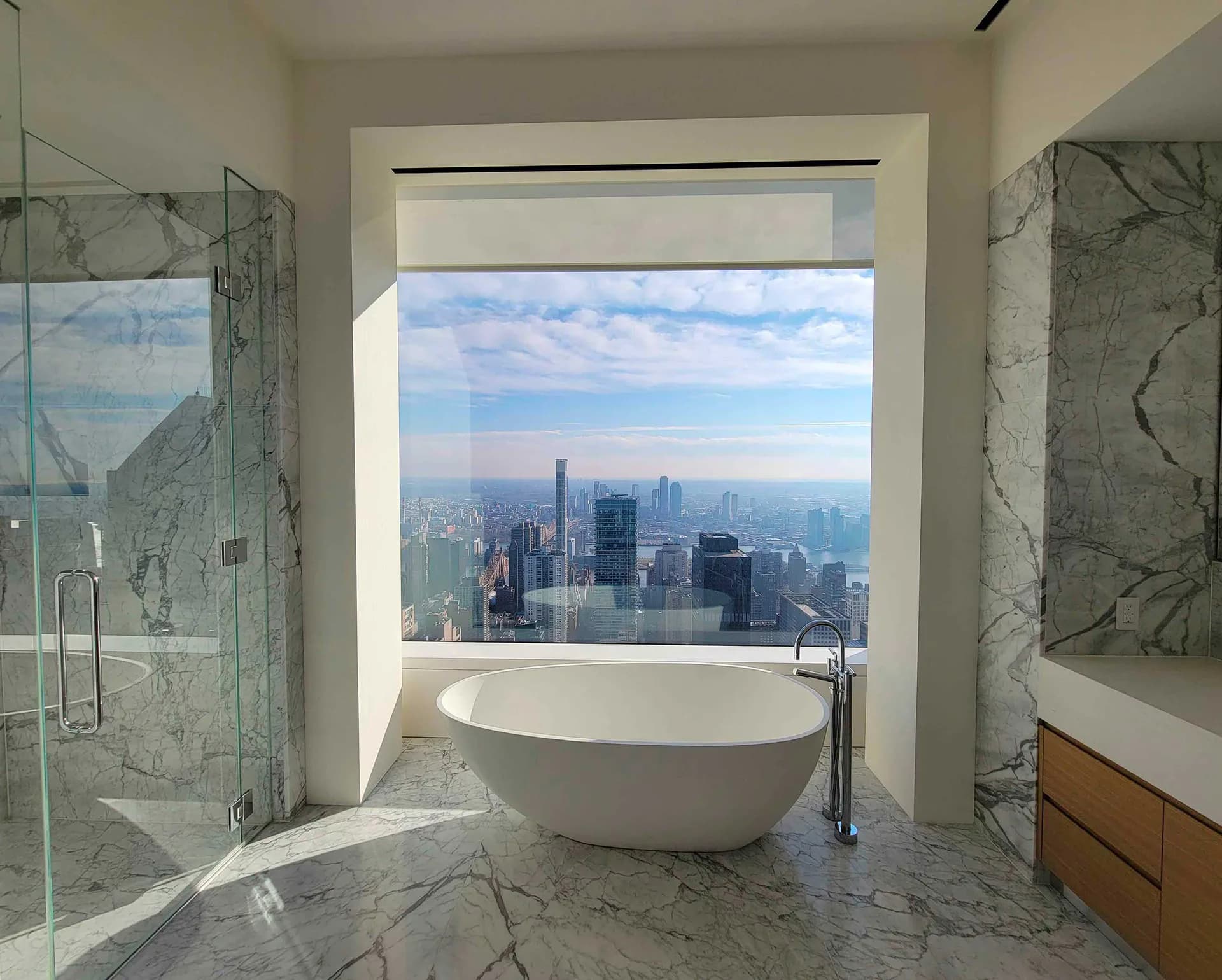 Luxurious marble bathroom with freestanding soaking tub positioned before floor-to-ceiling windows overlooking a city skyline, featuring geometric marble wall panels and glass shower enclosure by Atelier Premiere.