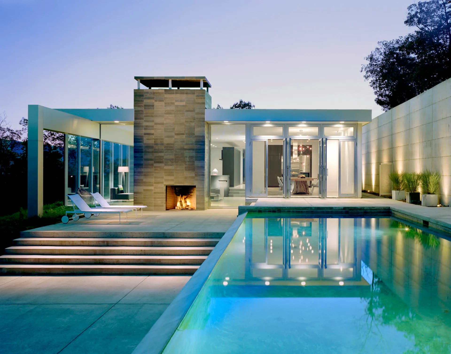 Modern residential architecture featuring a linear infinity pool, illuminated stone fireplace, and glass-walled pavilion with warm interior lighting at dusk by Audrey Matlock Architect.