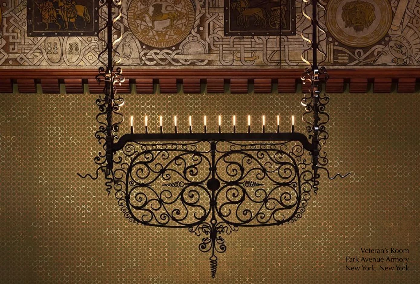 Ornate wrought iron decorative grille with scrollwork mounted on cream patterned wall below gilded ceiling detail by Aurora Lampworks.