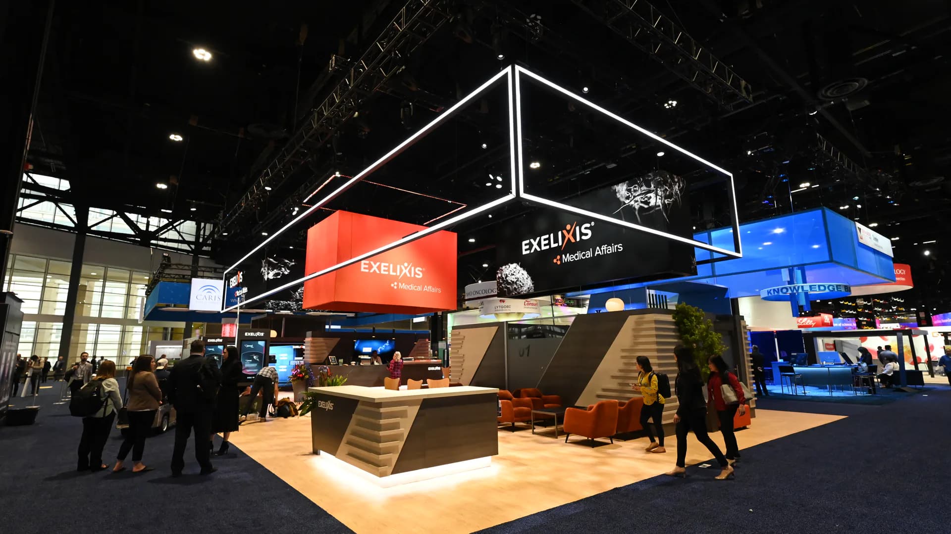 Modern trade show booth with angular black steel frame, orange and blue panel displays, wooden flooring and seating areas, surrounded by visitors in an indoor exhibition space by Available Light.