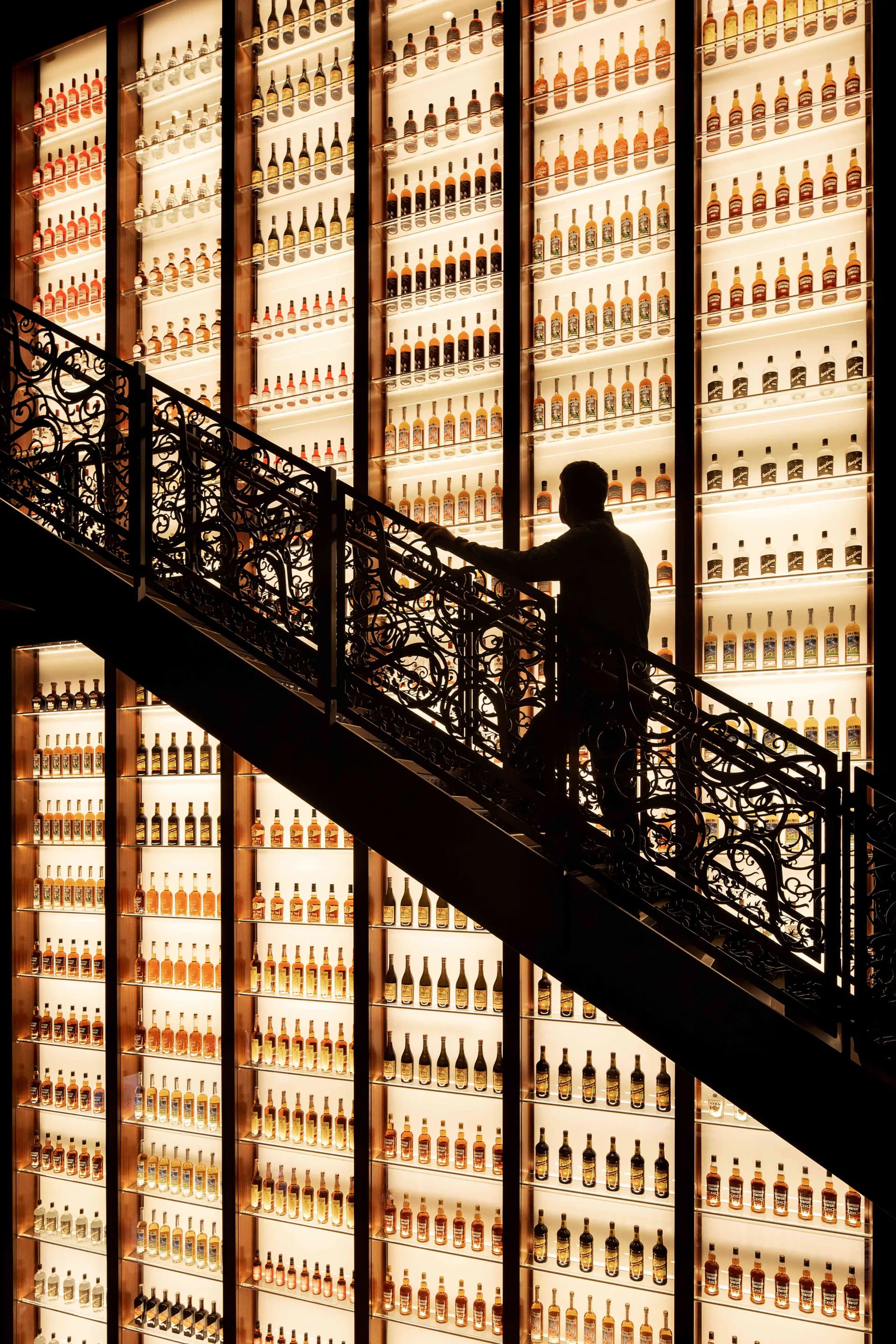 Silhouetted figure ascending a dramatic staircase with ornate metal railings, backdropped by an illuminated wall installation of hundreds of amber glass bottles by Available Light.