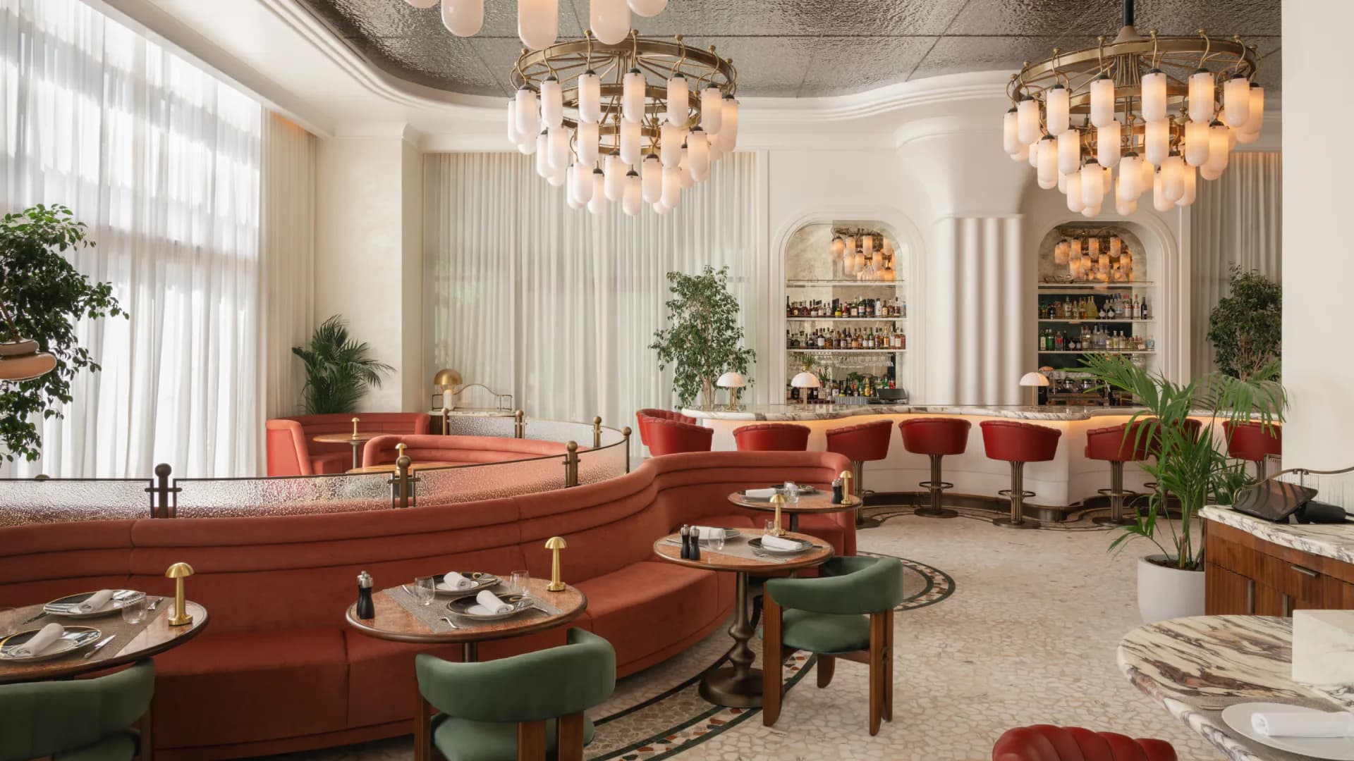 A luxurious lounge with curved red seating, green accent chairs, dual chandelier fixtures, polished columns, and abundant greenery throughout by AvroKO.