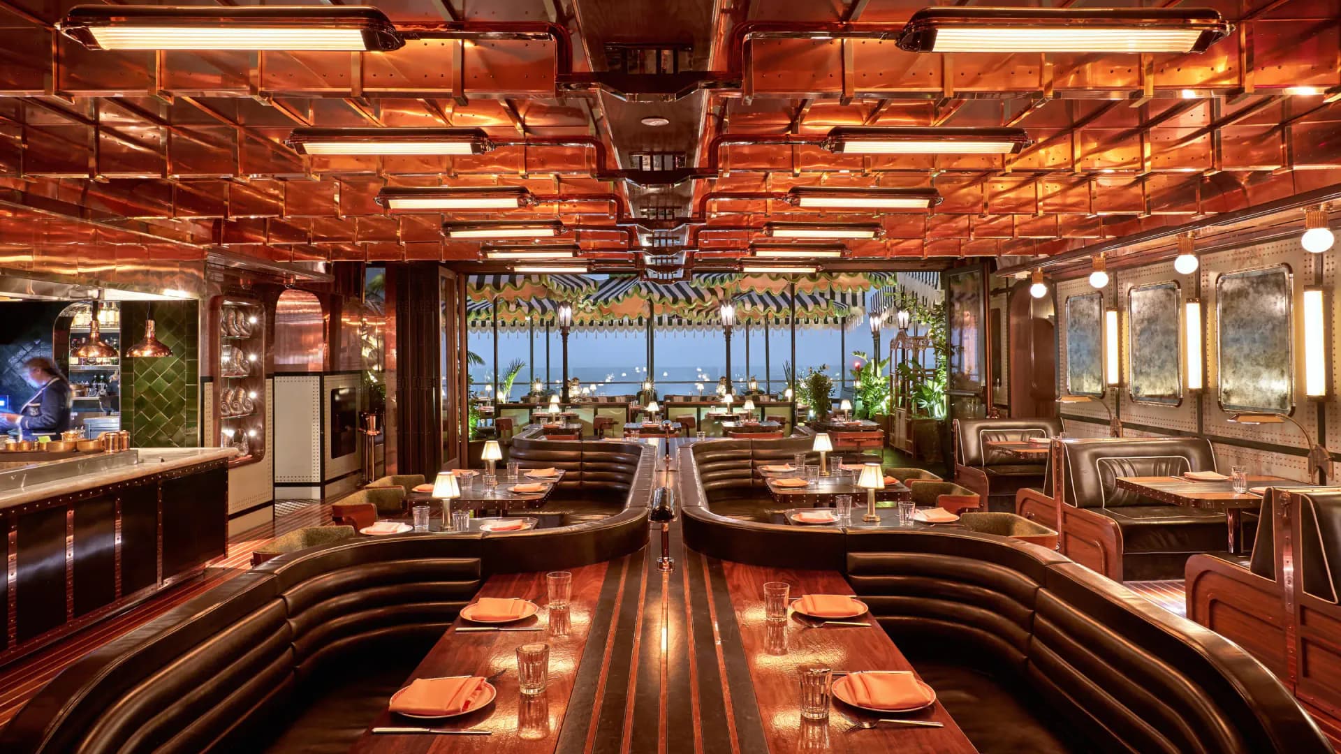 Warm-toned restaurant interior with copper ceiling elements, linear dining tables, and floor-to-ceiling windows overlooking a cityscape by AvroKO.