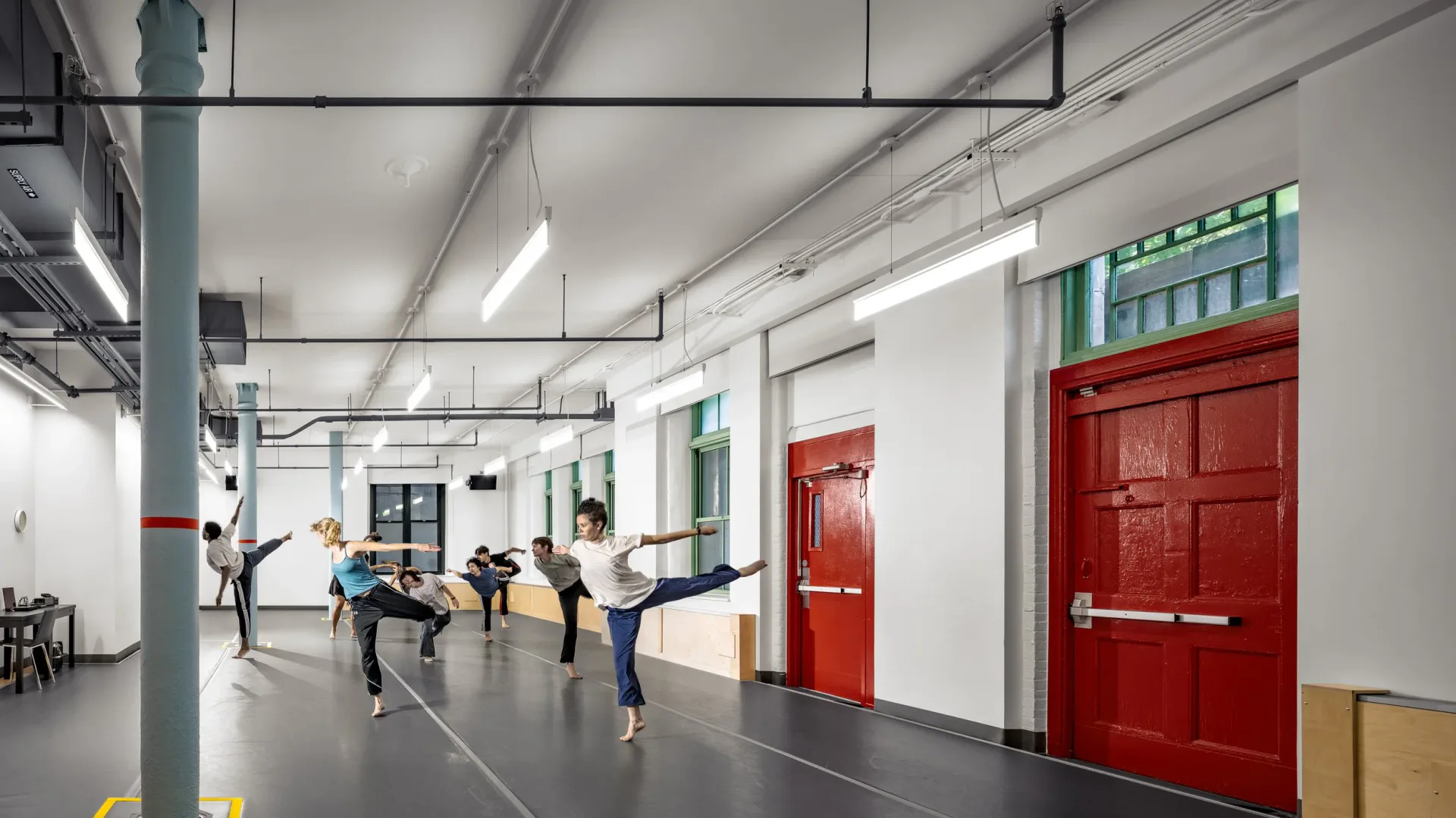 Industrial fitness studio with polished concrete floors, exposed ceiling systems, red doors, and green-framed windows; people in exercise class by AYON Studio Architecture and Preservation.