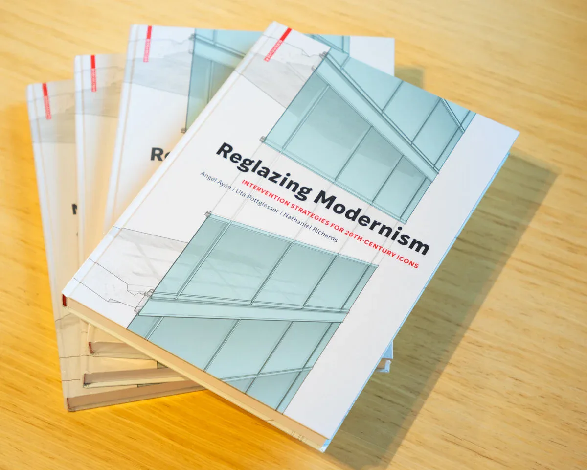 Stack of printed books titled 'Reglasing Modernism' with glass panel imagery on cover, displayed on wooden surface by AYON Studio Architecture and Preservation.