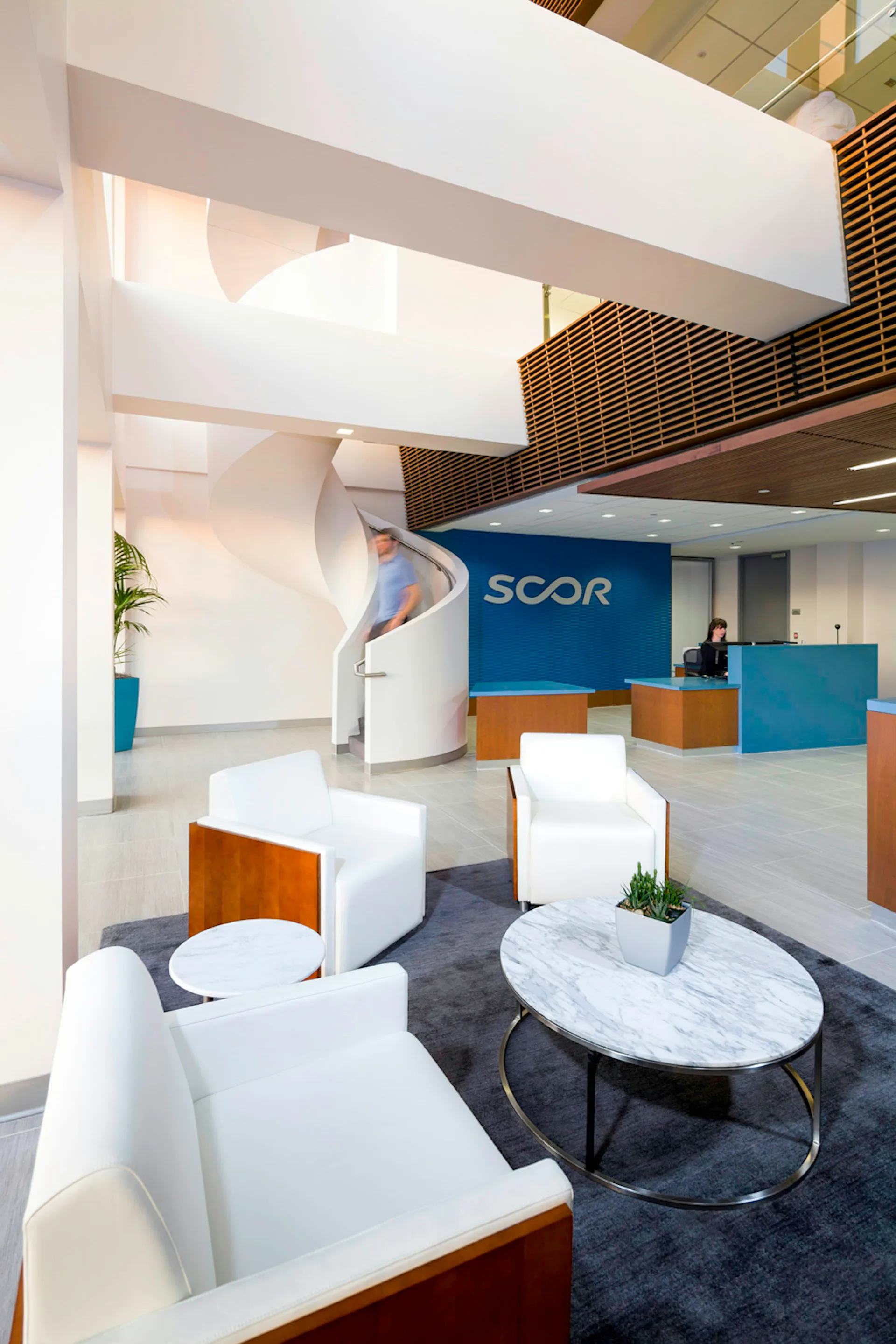Modern office lobby featuring white sculptural elements, blue SCOR signage, wooden accents, marble table, and multi-level white ceiling with natural light by BAM Creative.