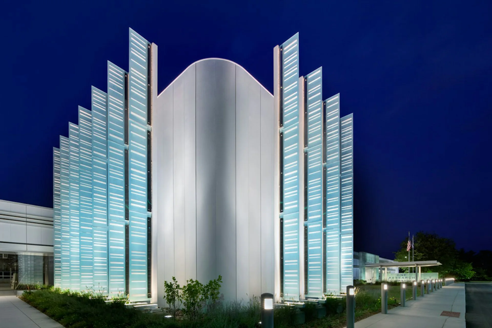 Illuminated white cylindrical tower with geometric glass wings at dusk, modern architectural rendering by BAM Creative.