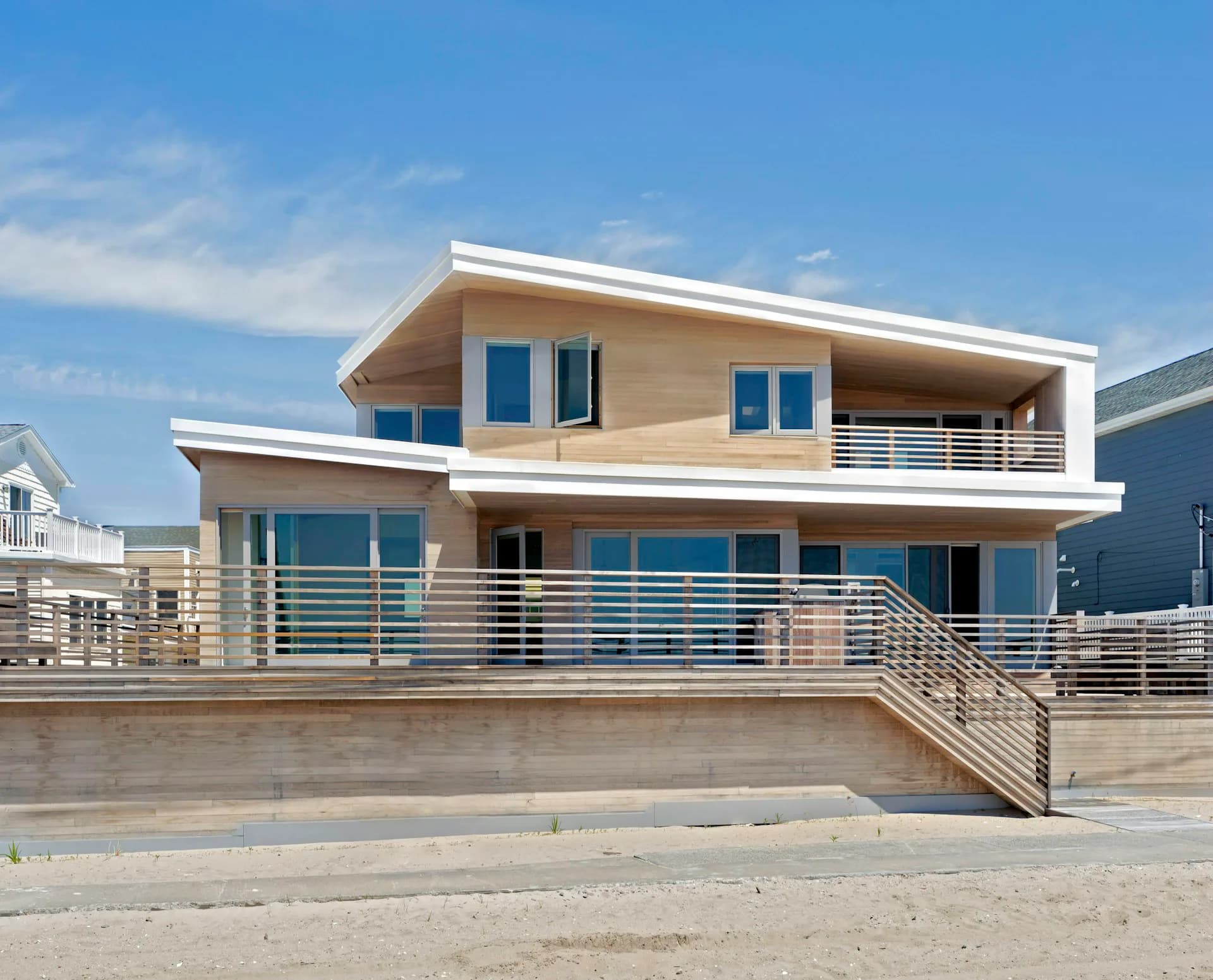 Modern two-story residential beach house with tan wood siding, blue windows, metal railings, and expansive deck overlooking sand by Barker Associates Architecture Office.