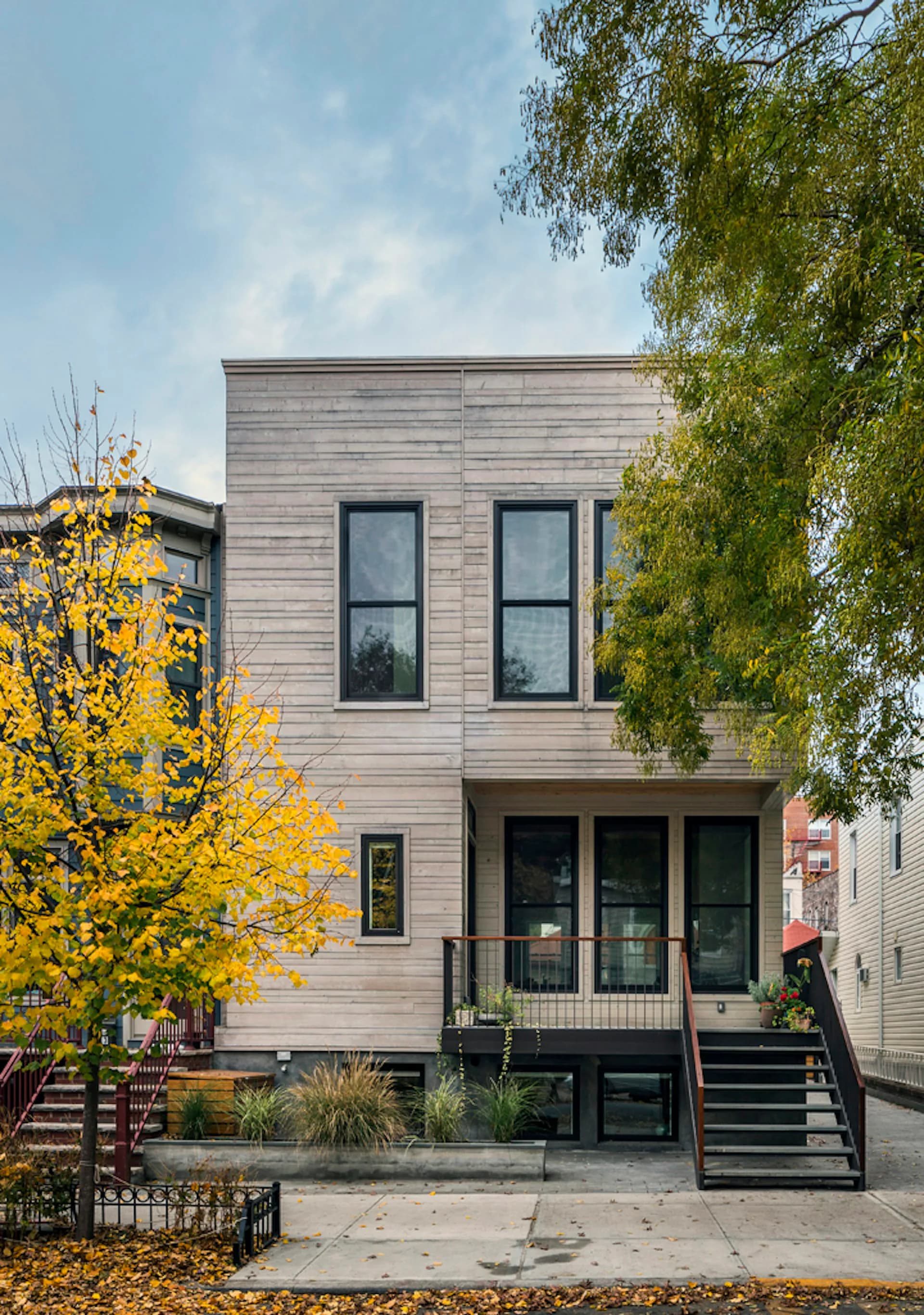 Modern residential building with natural wood siding, tall windows, and black metal staircase, surrounded by autumn foliage, by Barker Associates Architecture Office.