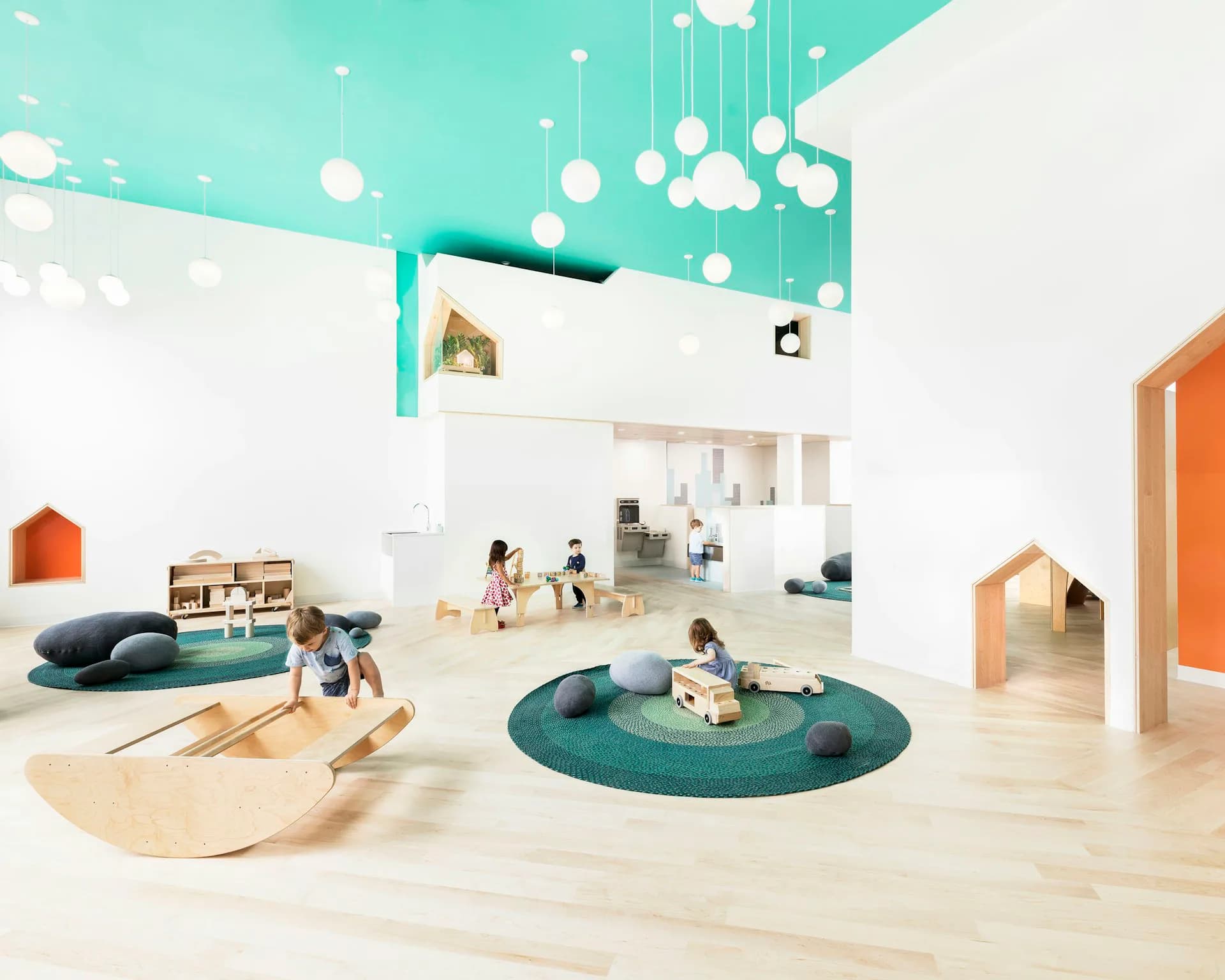 Bright early childhood education space with turquoise ceiling, light wood flooring, circular teal play areas, and orange accents, designed by Barker Associates Architecture Office.