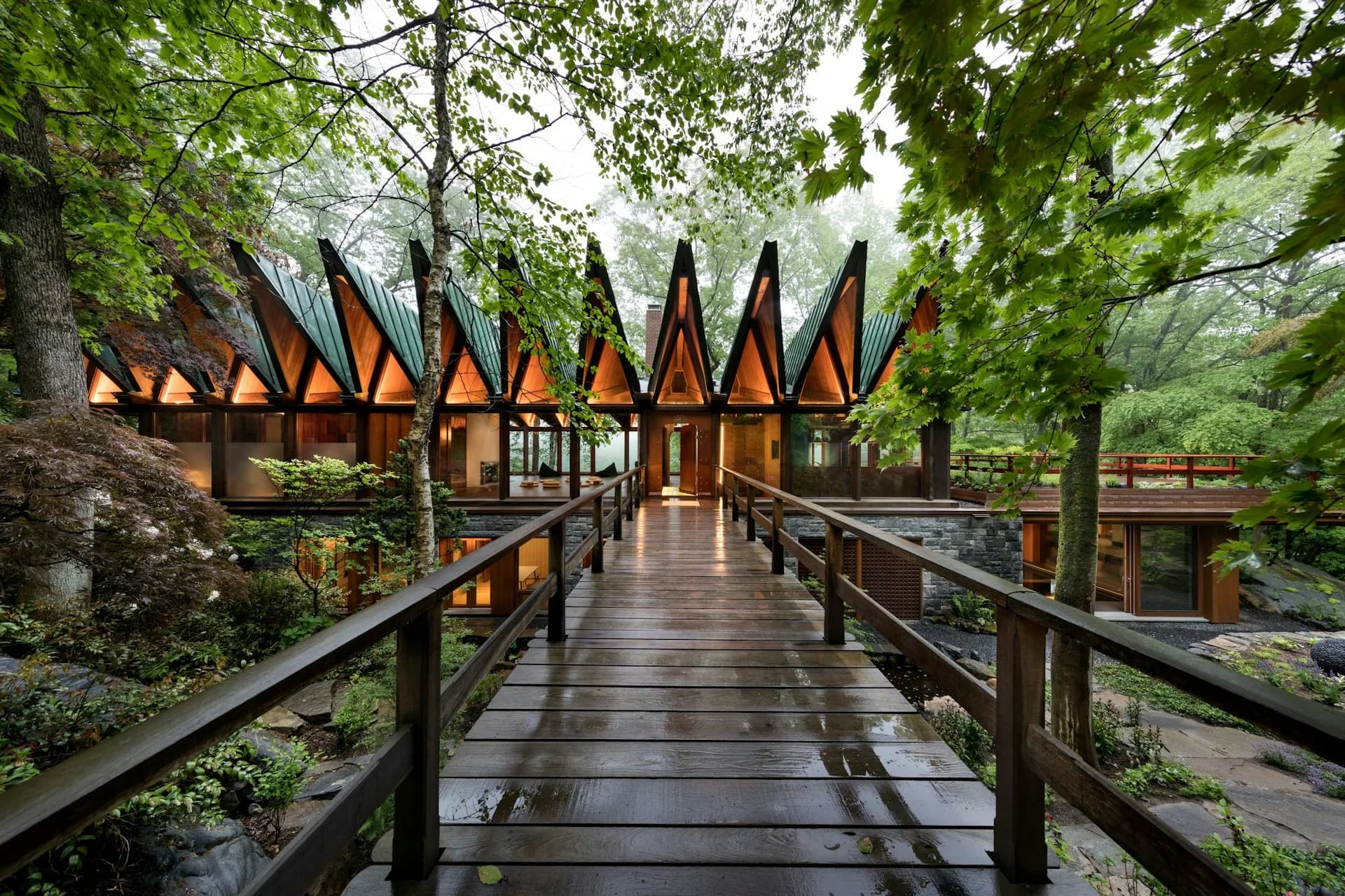 Modernist pavilion with distinctive peaked roof elements connected by wooden boardwalk through lush forest setting, designed by BarlisWedlick Architects LLC.