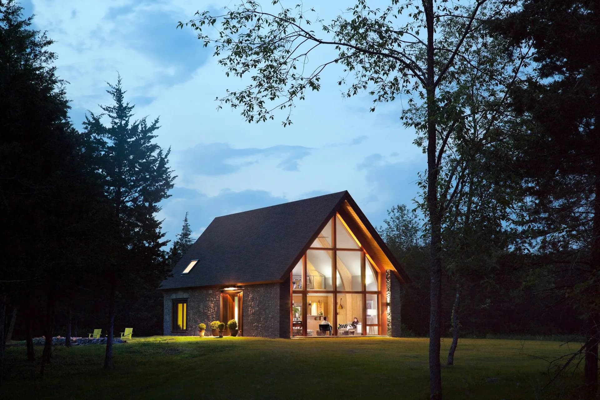 Modern residential house with dark exterior and large glazed gable end illuminated at dusk, set within a tree-lined clearing, by BarlisWedlick Architects LLC.