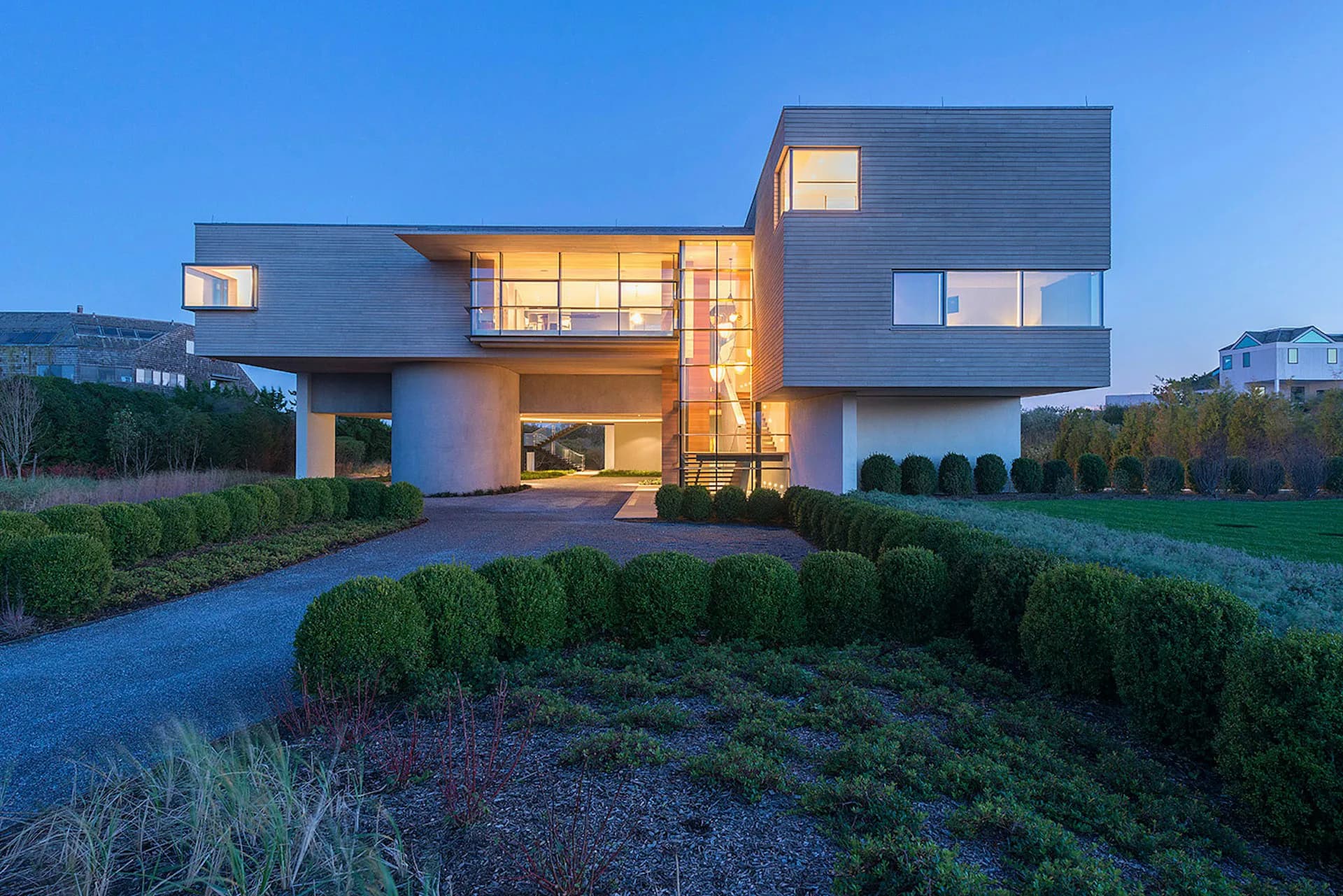 Modern residential architecture with geometric forms, illuminated interior spaces, and manicured landscape featuring boxwood hedges at dusk by Barnes Coy Architects.