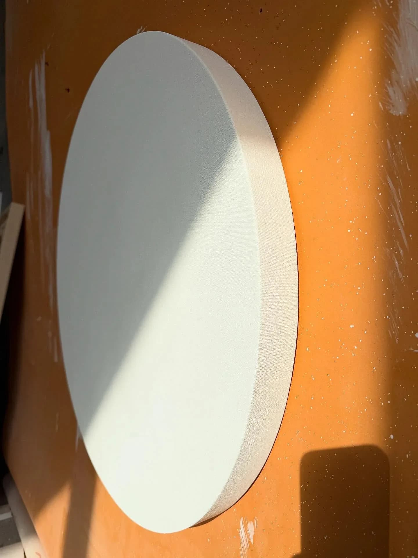 Minimalist oval wall-mounted light fixture with soft white glow against warm orange wall, casting geometric shadow, by Barronarts.
