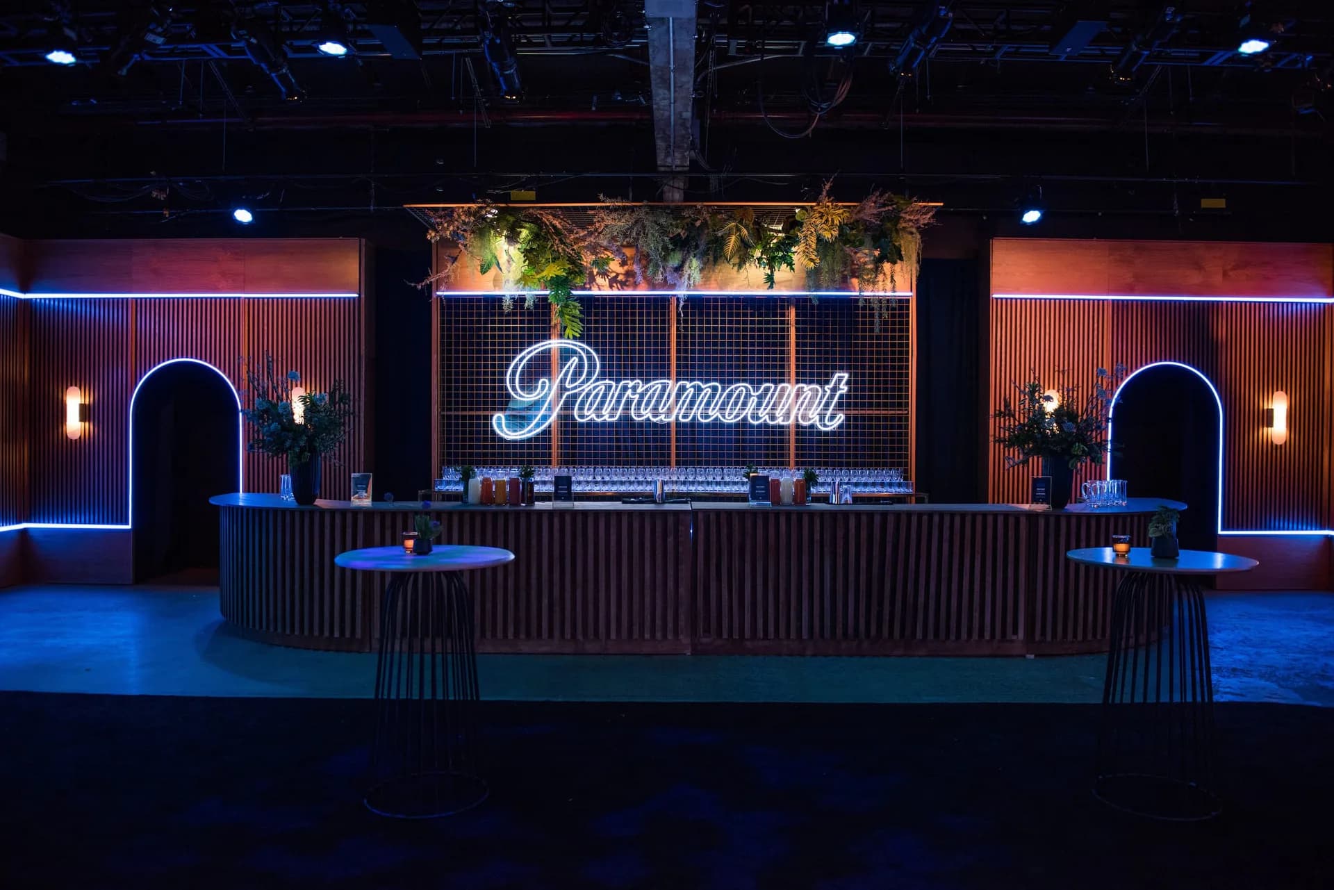 Modern event venue with blue ambient lighting, neon Paramount signage, geometric wall paneling, and minimalist bar counter by Bednark Studio.