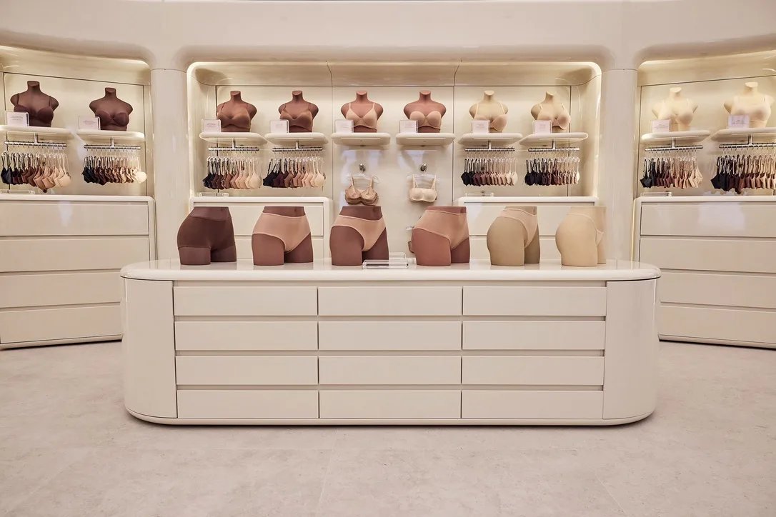 Minimalist retail display with curved white cabinetry, wall-mounted shelving showcasing clothing and accessories in warm earth tones by Bednark Studio.