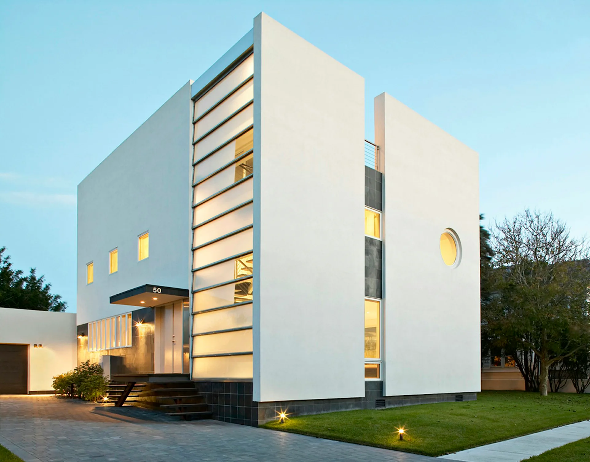 Modern white cubic residence with horizontal louver details, illuminated interior spaces, and manicured landscaping at dusk by Belmont Freeman Architects.