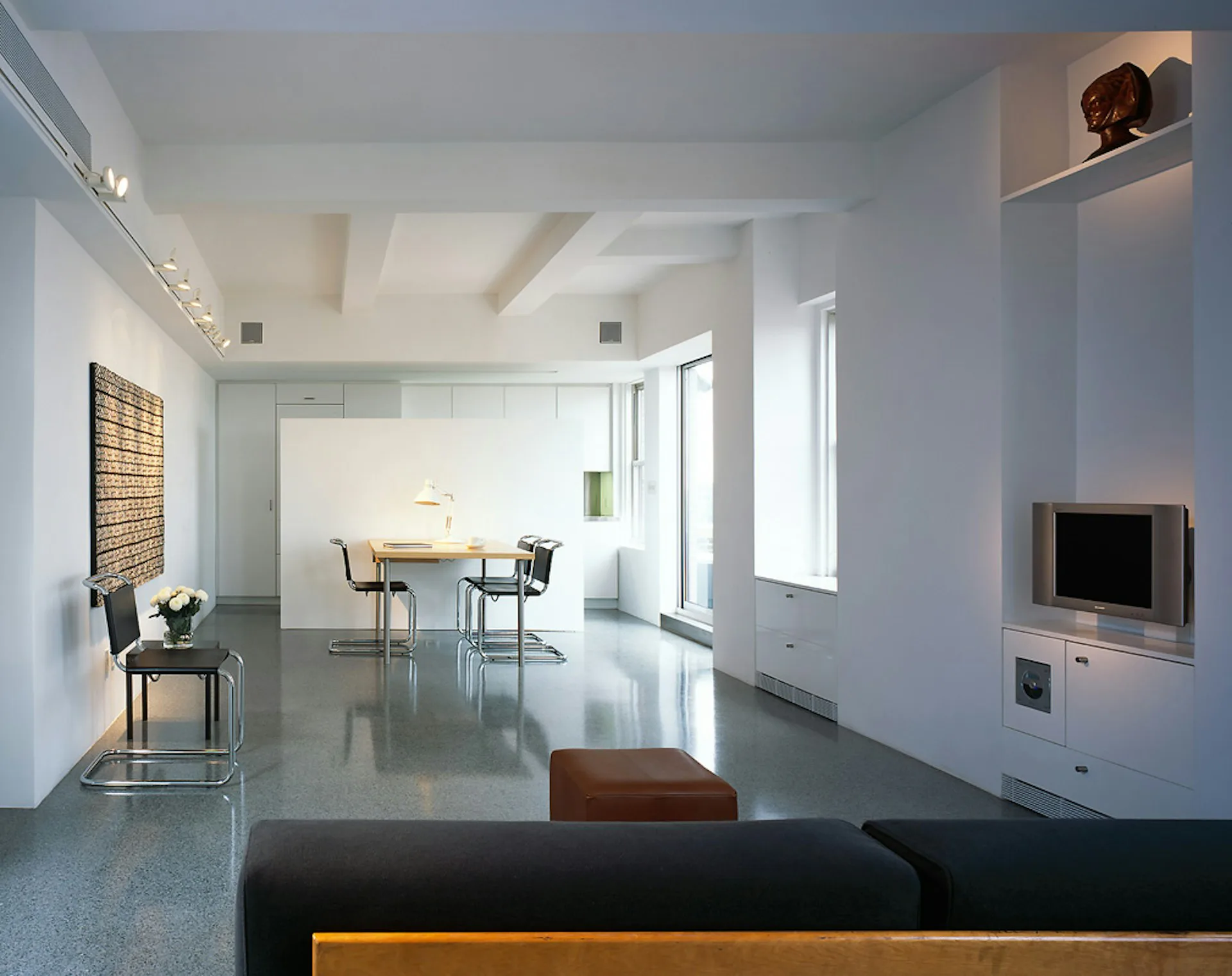 Minimalist loft interior with polished concrete floors, white walls, and natural light; dining area visible in background by Belmont Freeman Architects.