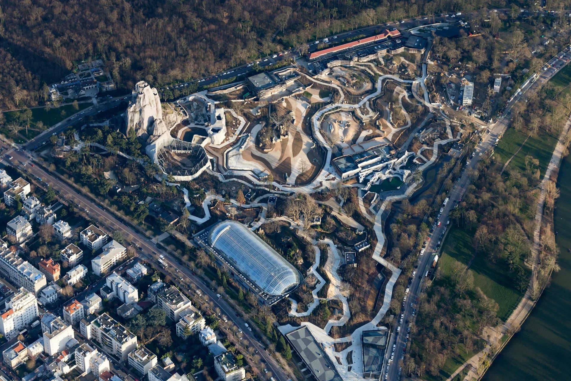 Aerial view of a large-scale landscape architecture project featuring serpentine pathways, terraced earthworks, and modernist structures nestled within forested terrain by Bernard Tschumi Architects.