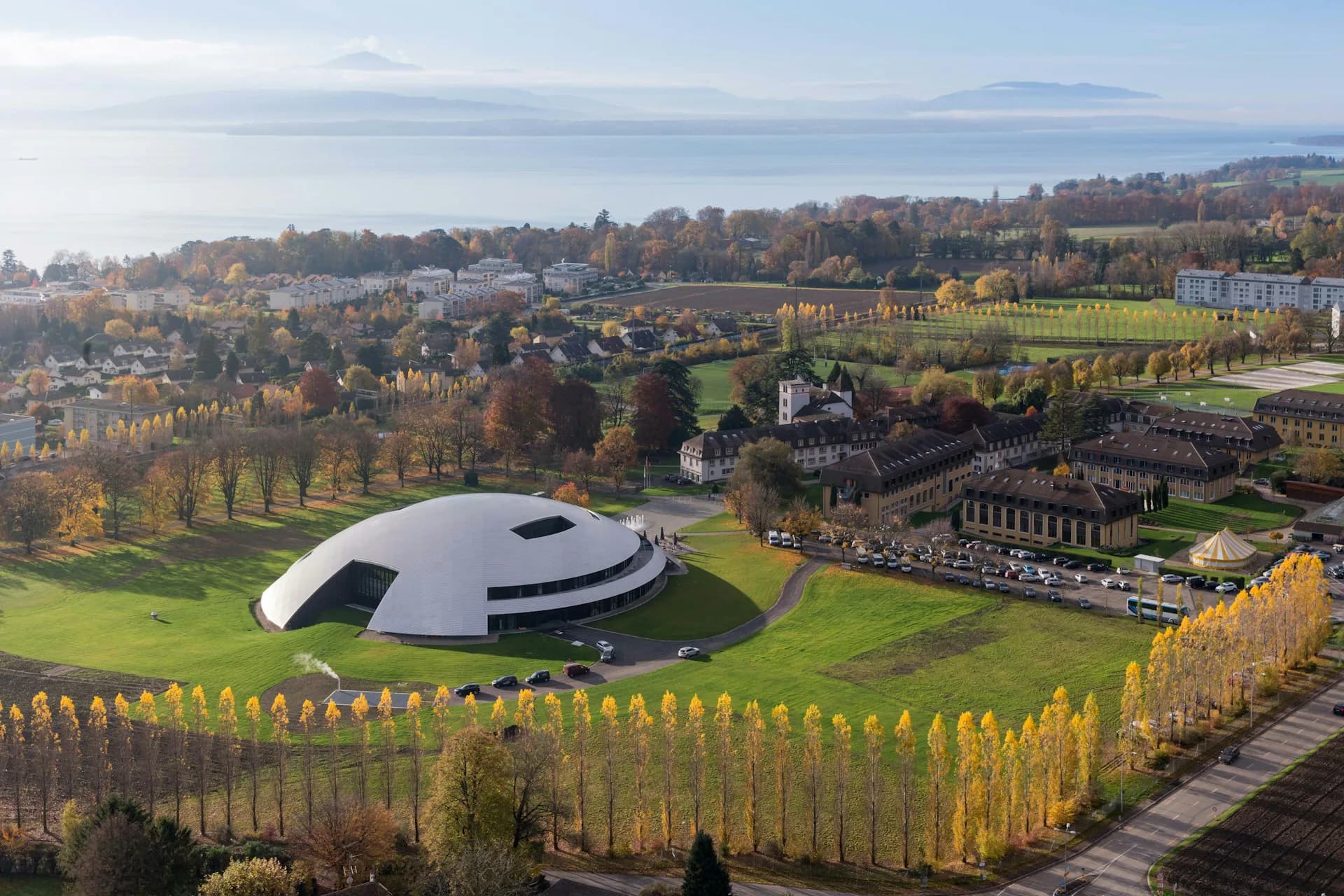 Aerial view of a white disc-shaped modern building surrounded by green landscape and golden autumn trees, with residential areas visible in the distance, by Bernard Tschumi Architects.