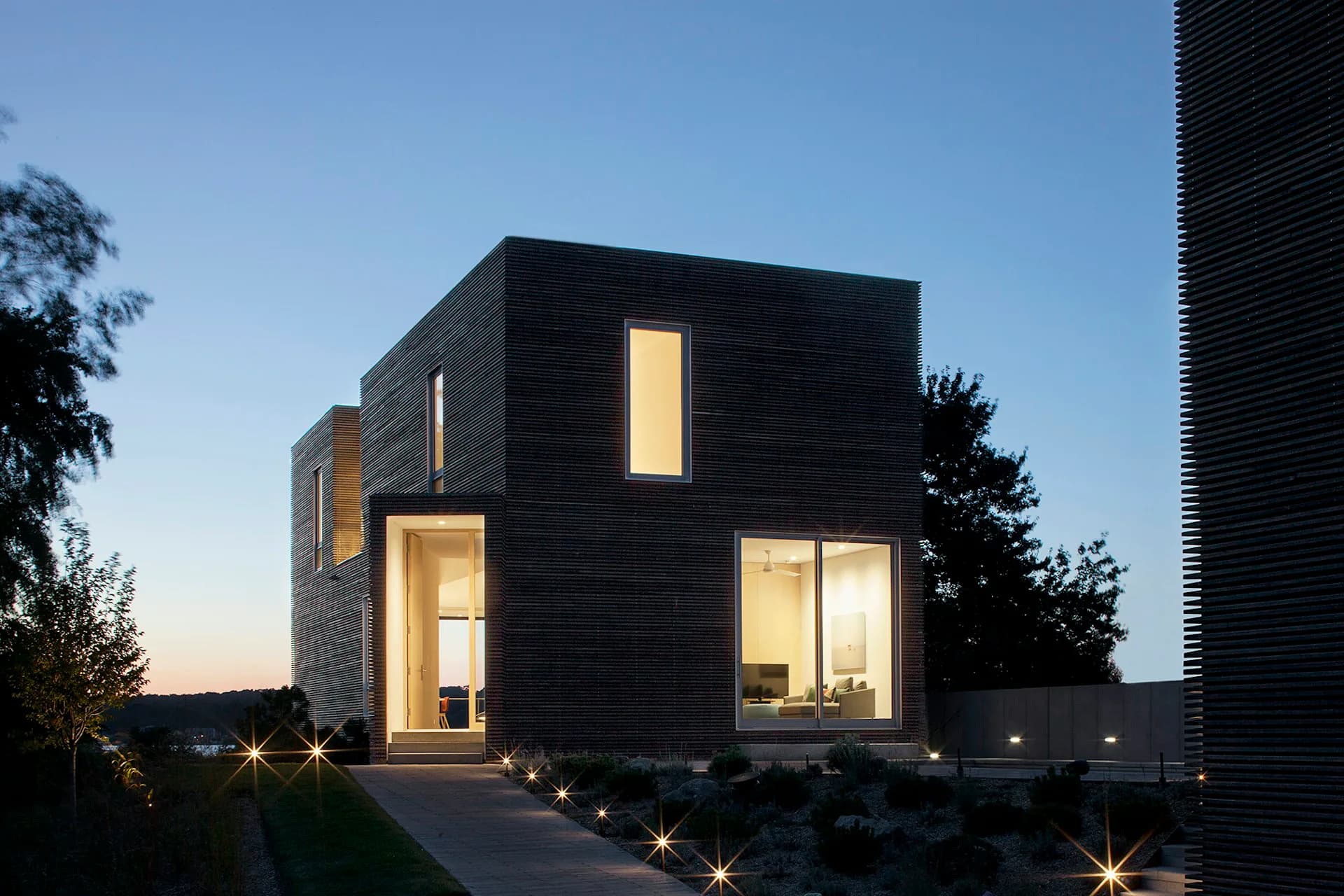 Modern cubic black residence with warm interior lighting, ground-level uplighting, and minimalist landscaping at dusk by Bernheimer Architecture PLLC.