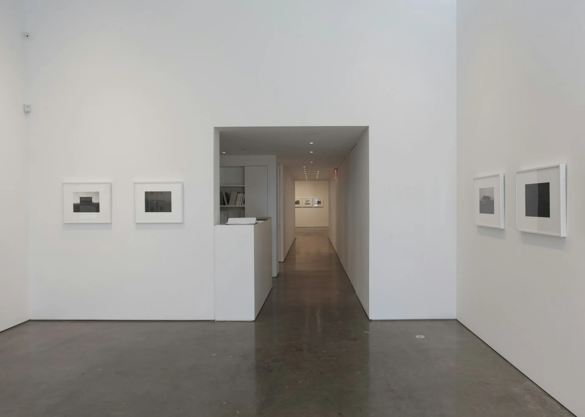 Minimalist gallery space with polished concrete floors, white walls, and framed artworks, featuring a central hallway with warm wooden flooring, designed by Bernheimer Architecture PLLC.