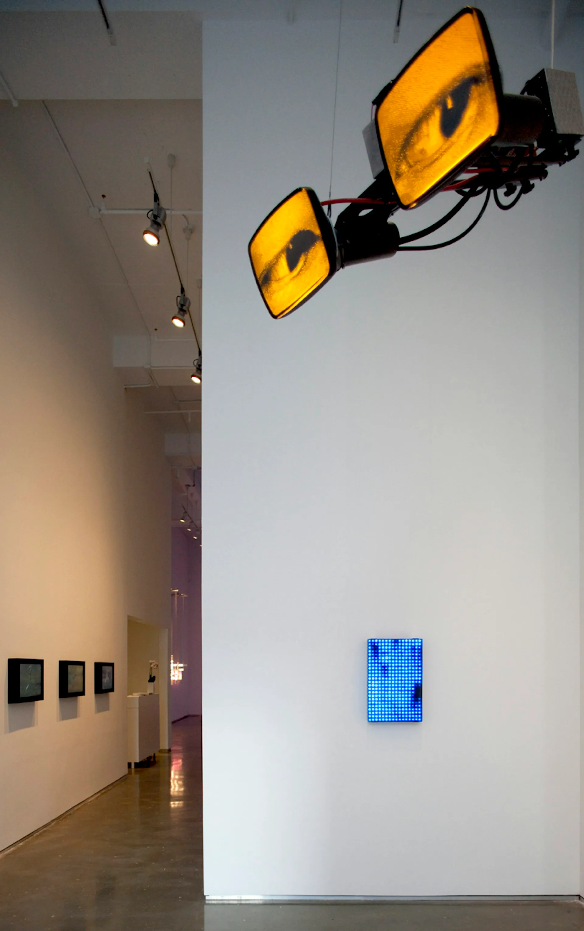 Contemporary gallery space with polished concrete floors, white walls, and suspended yellow sculptural lighting fixtures alongside a blue LED panel installation by Bernheimer Architecture PLLC.