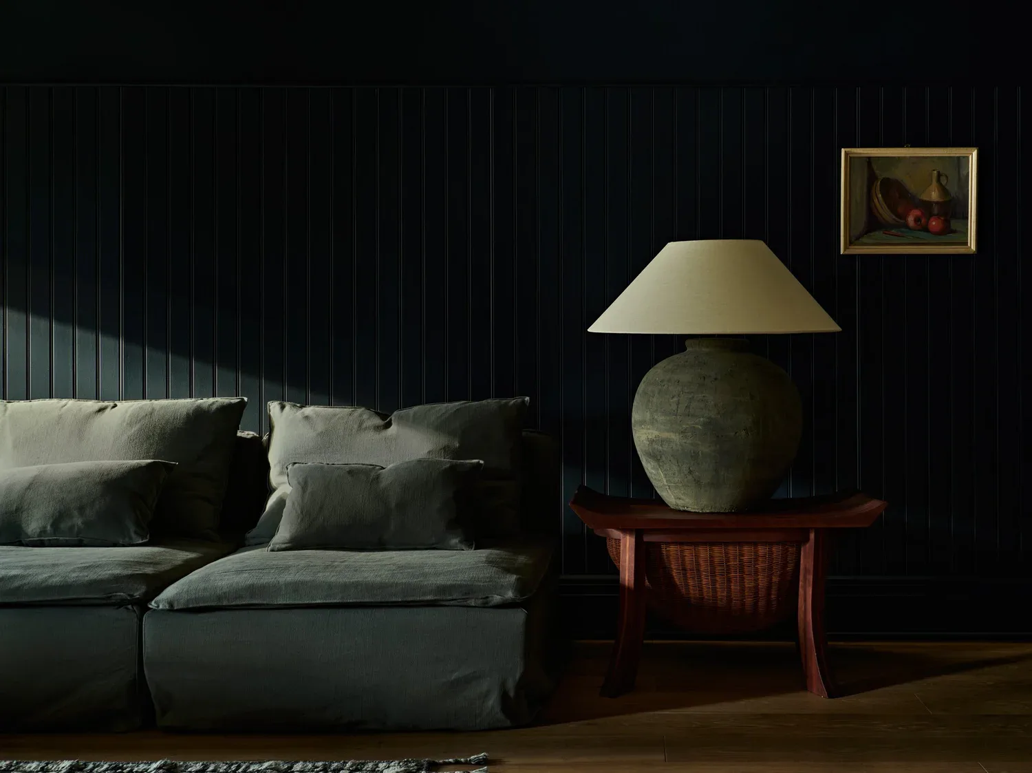 Dark modern bedroom with grey upholstered bed, wooden side table with brass lamp, red chair, and framed artwork, designed by Bespoke Only.