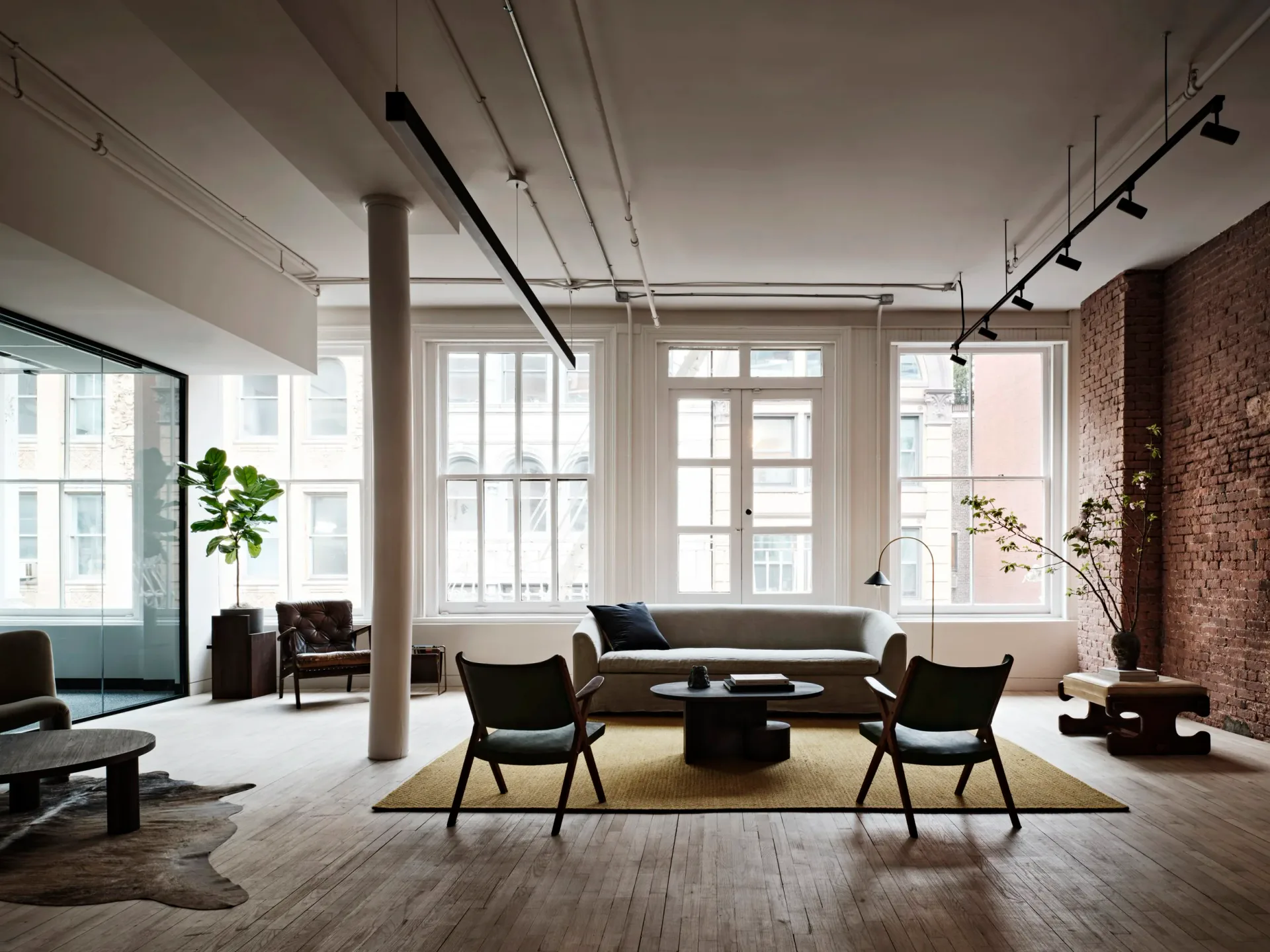 Modern loft interior with exposed brick, concrete columns, and floor-to-ceiling windows featuring a gray sofa, mid-century chairs, and natural wood flooring by Bespoke Only.