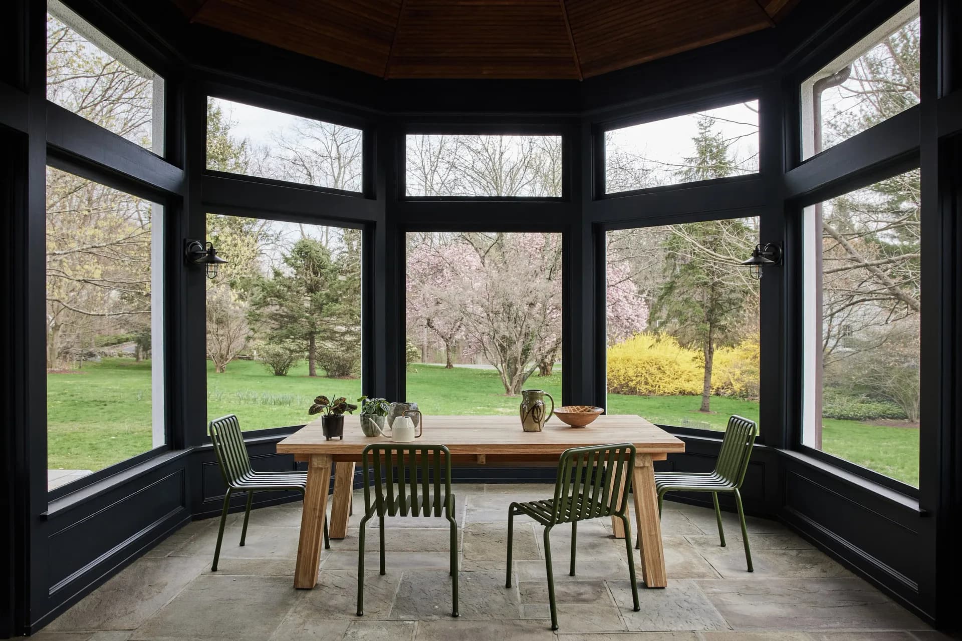 Modern sunroom with curved black-framed glass walls, wooden dining table, green chairs, and expansive views of manicured grounds and trees by Bespoke Only.