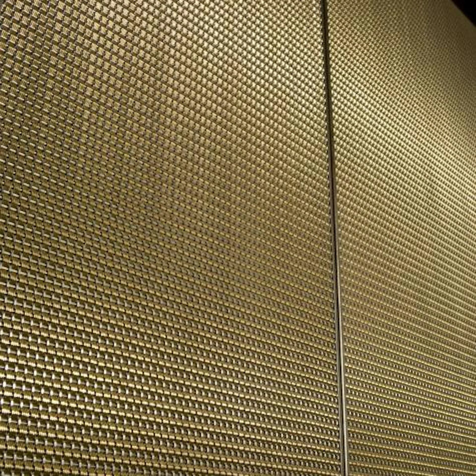 Close-up detail of a textured wall surface with woven metallic mesh or perforated metal paneling in gold tones, featuring a clean vertical seam, by BG Studio International Inc.