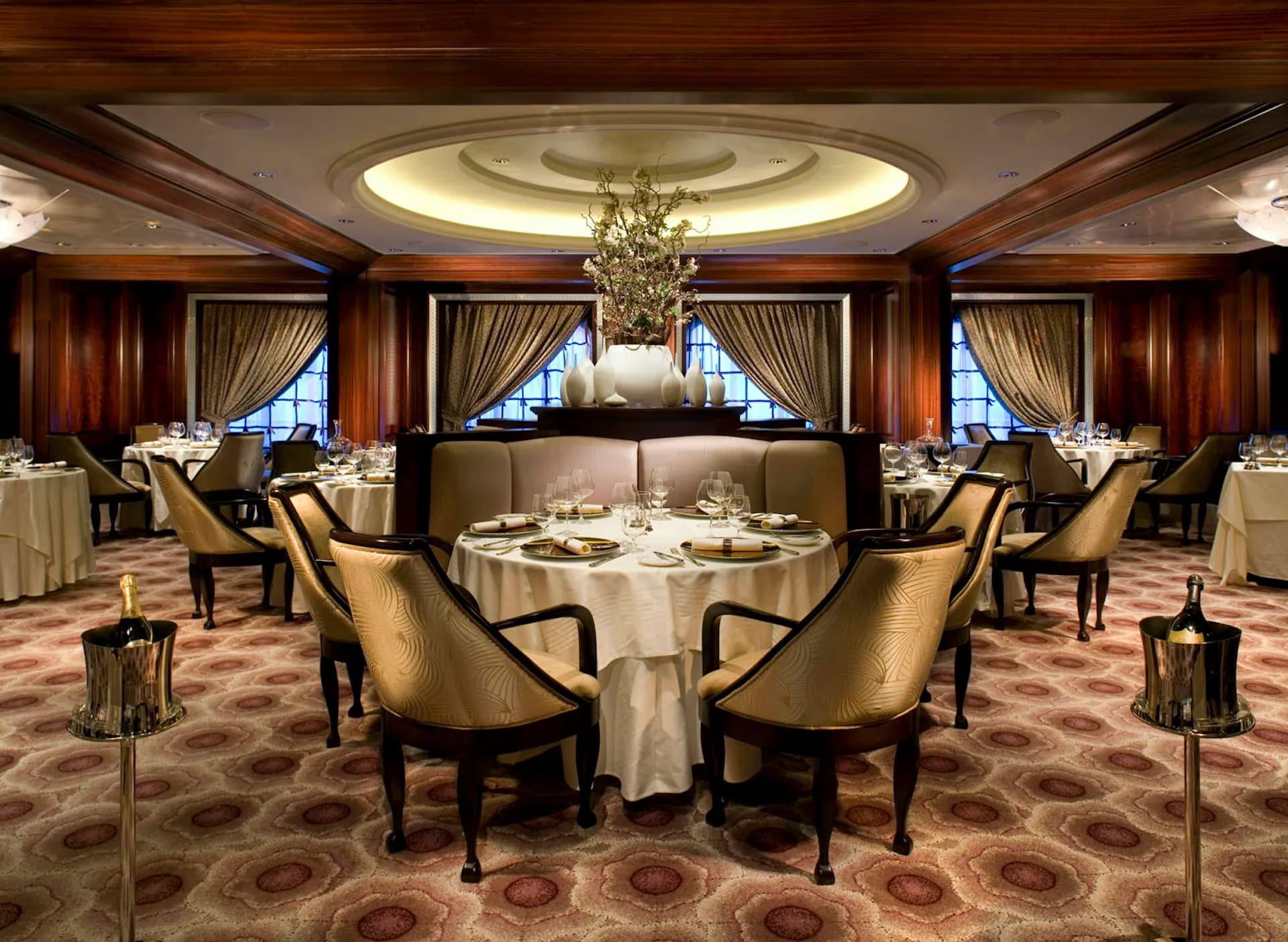 Elegant fine-dining restaurant with mahogany wood paneling, crystal chandelier, gold upholstered chairs, and round white-clothed tables by BG Studio International Inc.