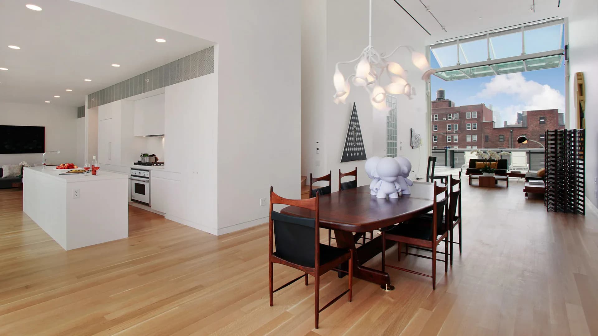 Modern loft dining and kitchen space with white cabinetry, wood flooring, pendant lighting, and floor-to-ceiling windows overlooking a city skyline by Bien Hecho.
