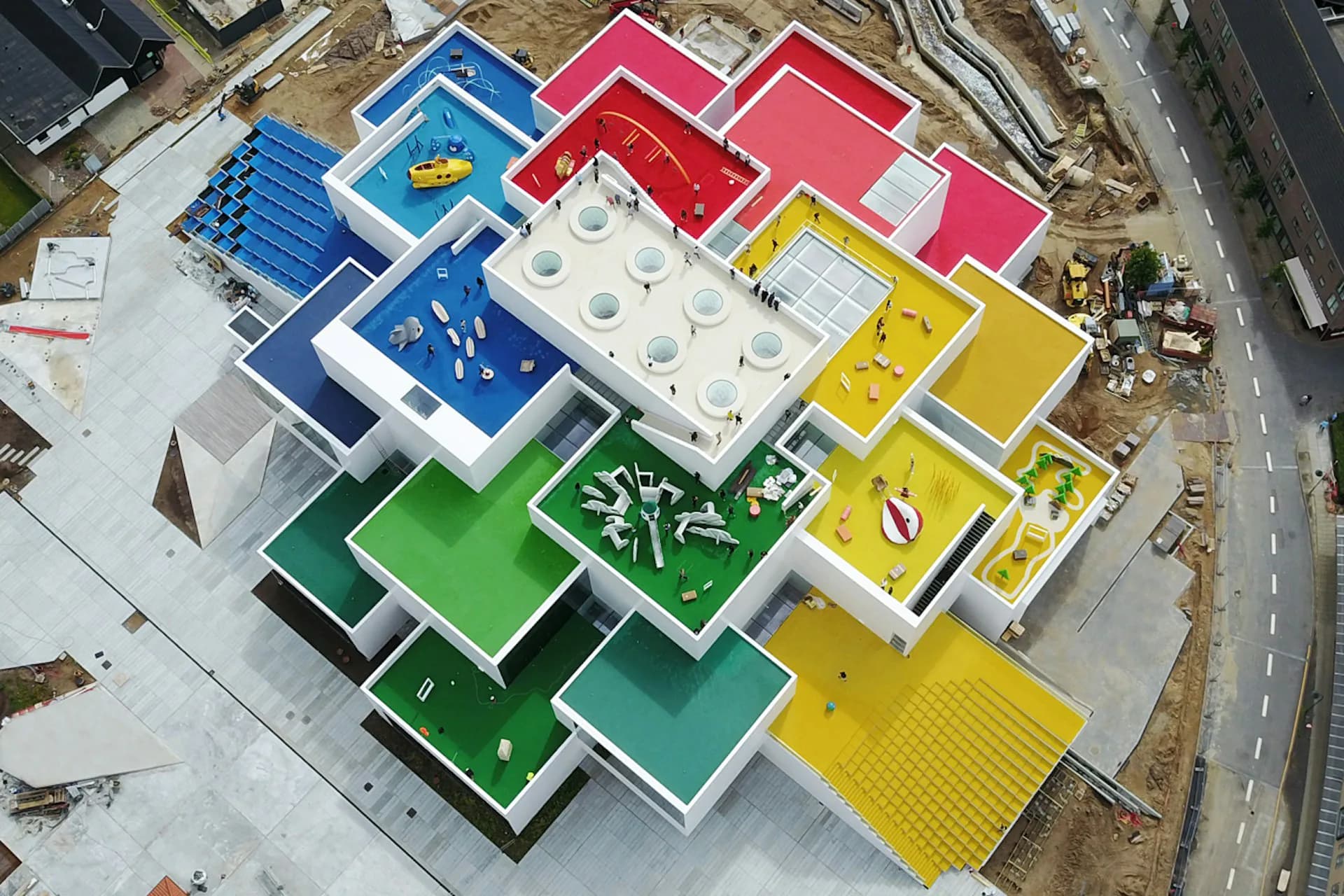 Aerial view of a colorful modular playground structure with blue, red, yellow, and green sections featuring various play equipment and climbing features, designed by BIG - Bjarke Ingels Group.