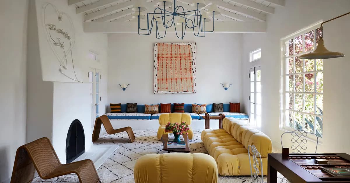 Bright white living room with golden-yellow curved seating, woven wall hanging, blue-toned cushions, and sculptural furniture by Billy Cotton Studio.