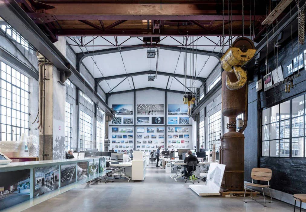 Spacious industrial loft with exposed steel trusses, large windows, concrete floors, and scattered sculptural objects and furniture throughout, designed by Bjarke Ingels Group — BIG NYC.