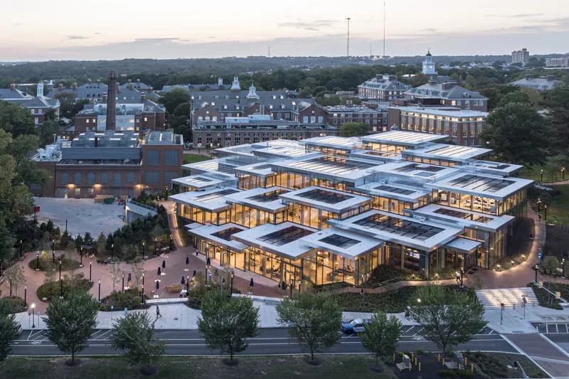 Aerial view of a contemporary institutional building with warm-lit glass and wood facades arranged in modular blocks around courtyards, set in a historic town landscape at dusk, by Bjarke Ingels Group — BIG NYC.