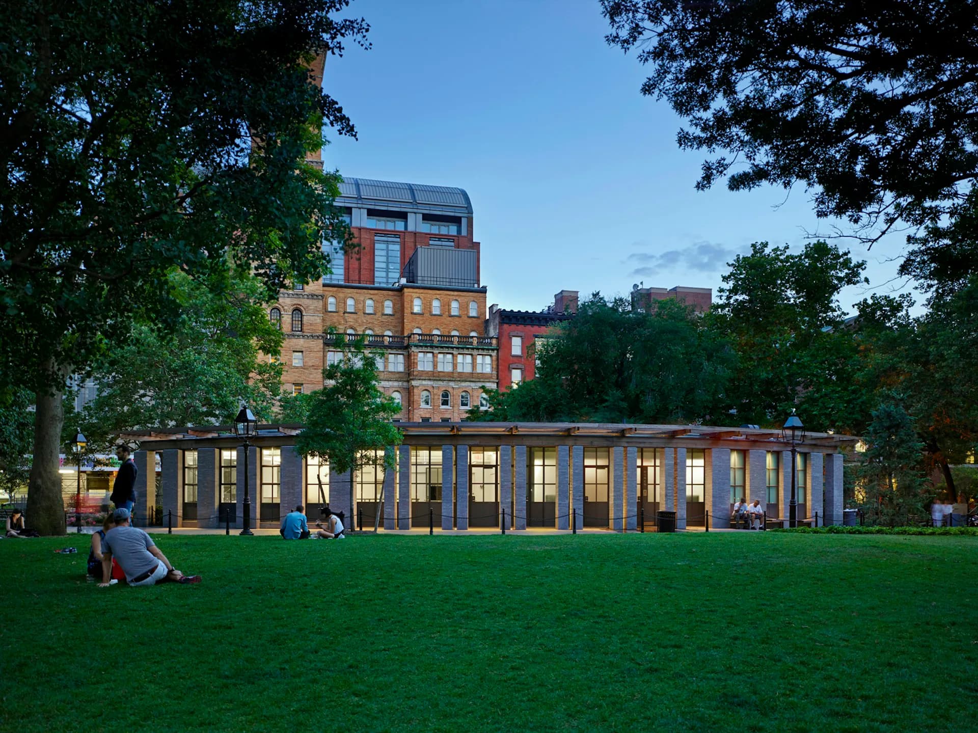 Modern pavilion with glass walls and warm interior lighting sits on a grassy park lawn surrounded by mature trees and historic brick buildings by BKSK Architects.