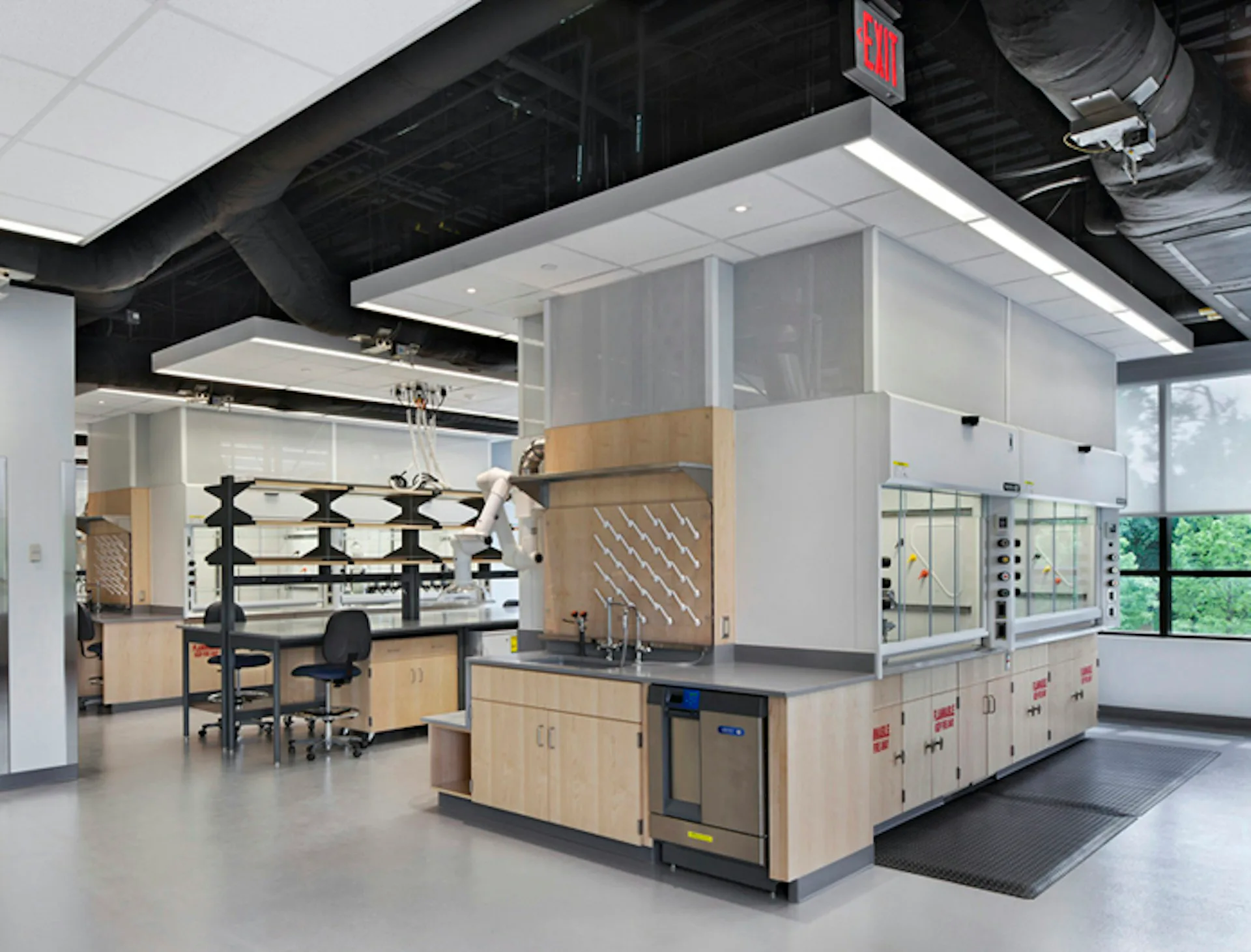 Modern commercial kitchen with light wood cabinetry, white upper units, and a central island featuring stainless steel appliances, designed by Blair + Mui Dowd Architects.