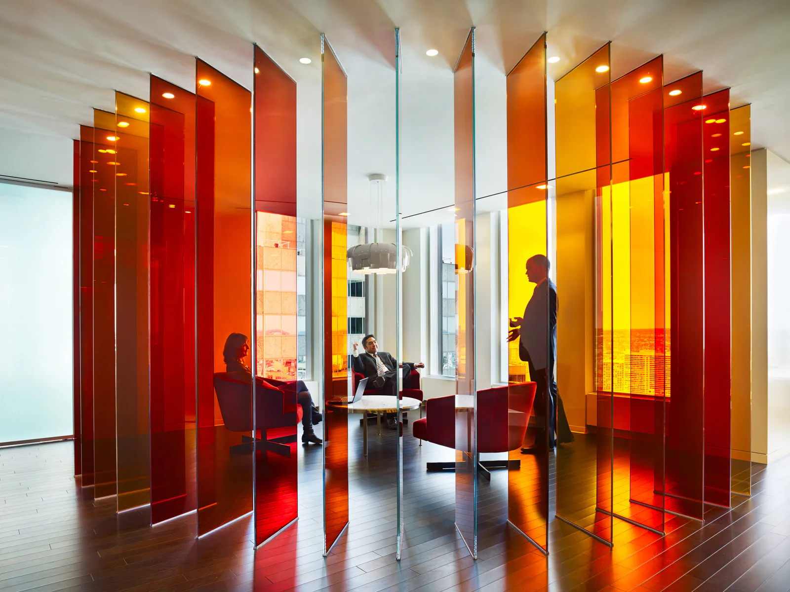 Modern office interior with layered translucent red and orange glass partitions creating a dynamic spatial composition, featuring silhouetted figures and warm ambient lighting by Bliss Fasman.