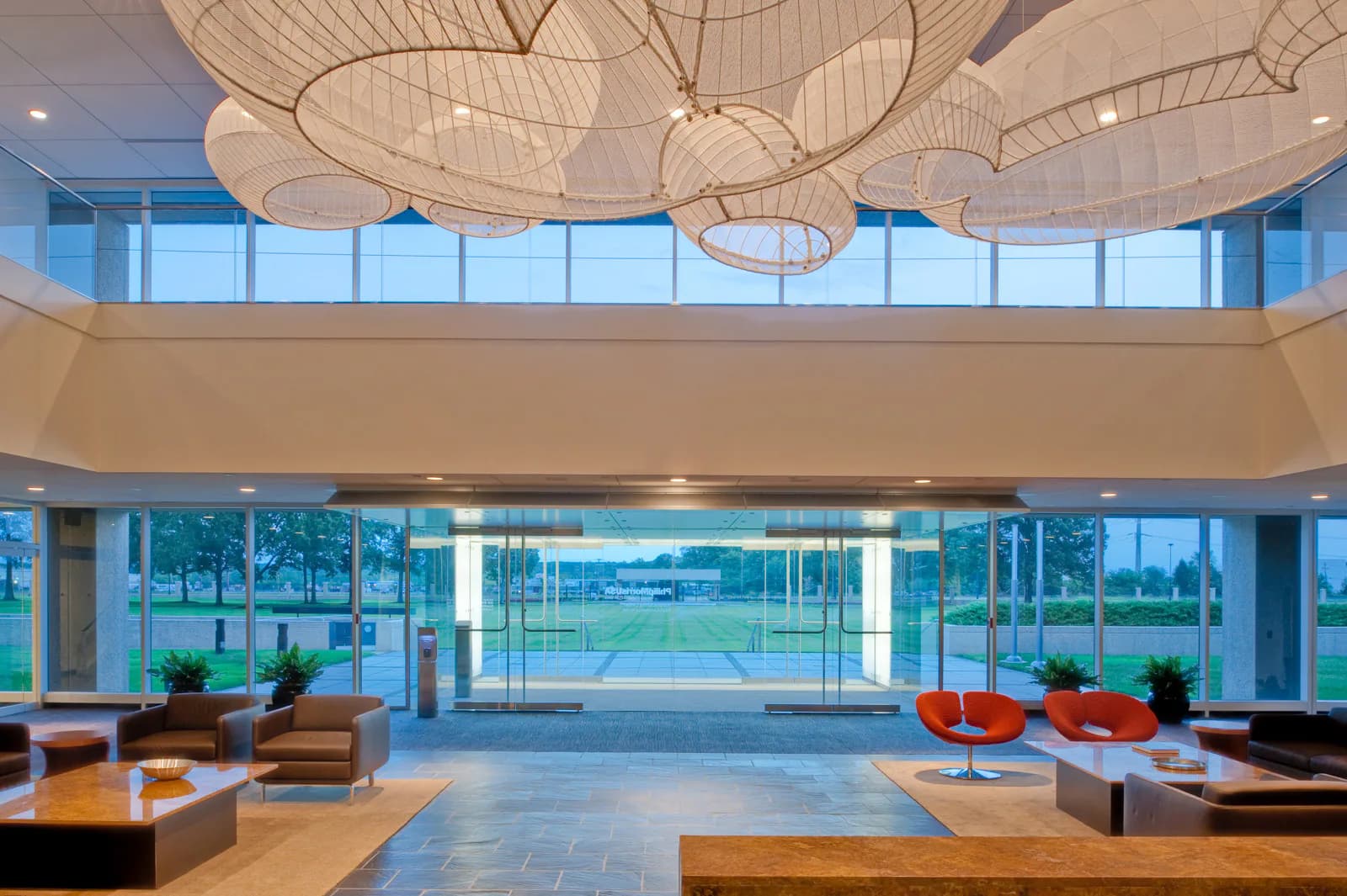 Modern interior with floor-to-ceiling windows overlooking a golf course, featuring sculptural pendant lighting, wood furnishings, and minimalist seating areas by Bliss Fasman.