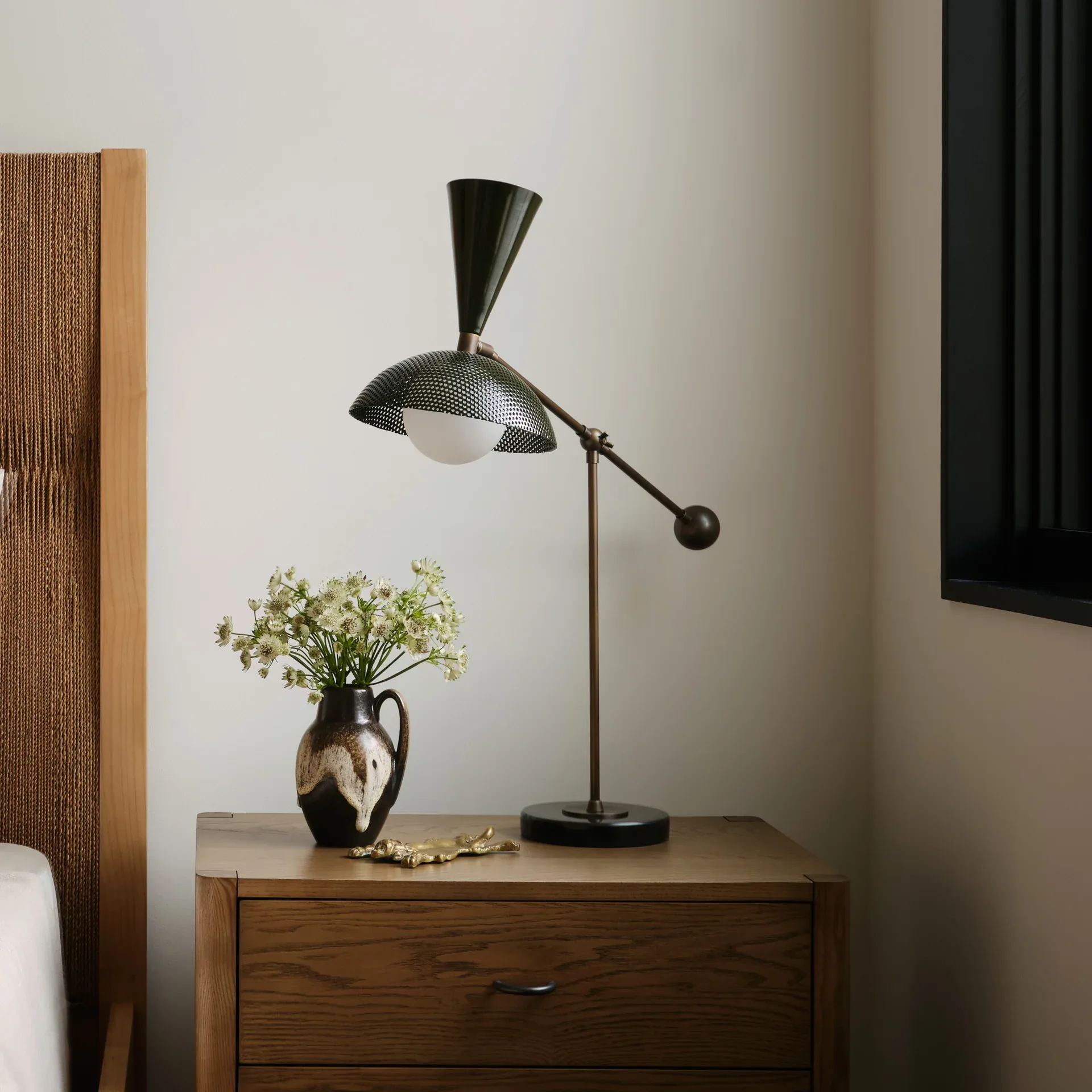 Minimalist bedside vignette with a sculptural black task lamp, ceramic pitcher with greenery, and warm wood nightstand against neutral walls by Blueprint Lighting.
