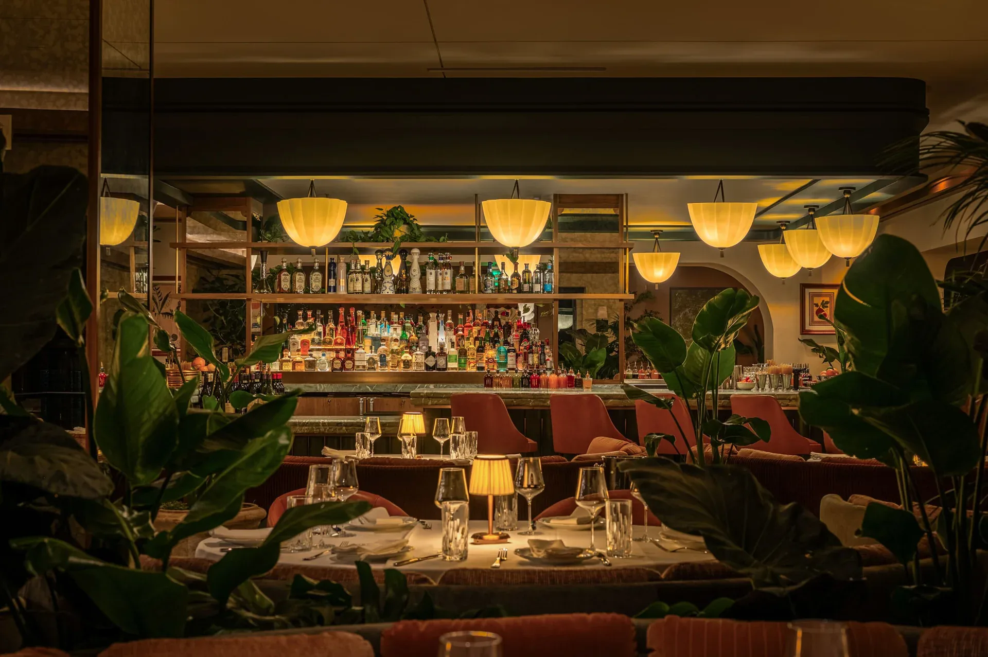 Elegant restaurant dining room with warm amber pendant lighting, dark wood furnishings, plant accents, and illuminated bar backdrop by Blueprint Lighting.