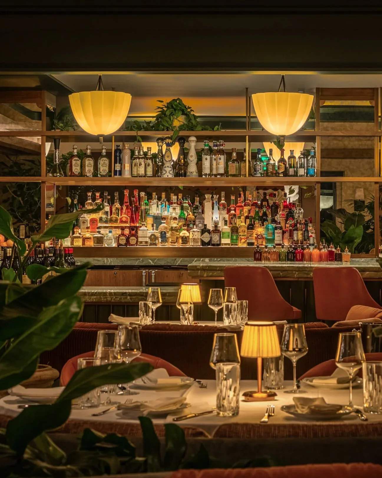 Upscale bar and dining space with warm amber pendant lighting, dark wood shelving stocked with bottles, burgundy seating, and lush green plants throughout, designed by Blueprint Lighting.