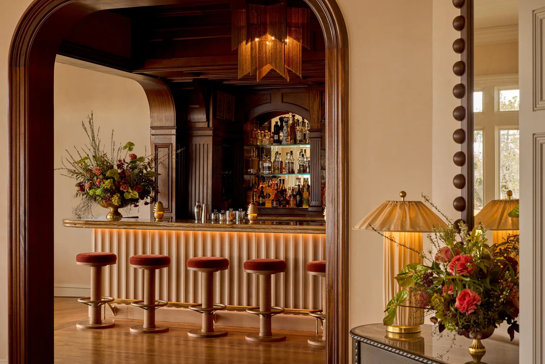 Elegant bar interior with warm wood tones, burgundy leather stools, striped paneling, and brass pendant lighting fixtures showcasing sophisticated ambient and task lighting by BOLD (Brian Orter Lighting Design).