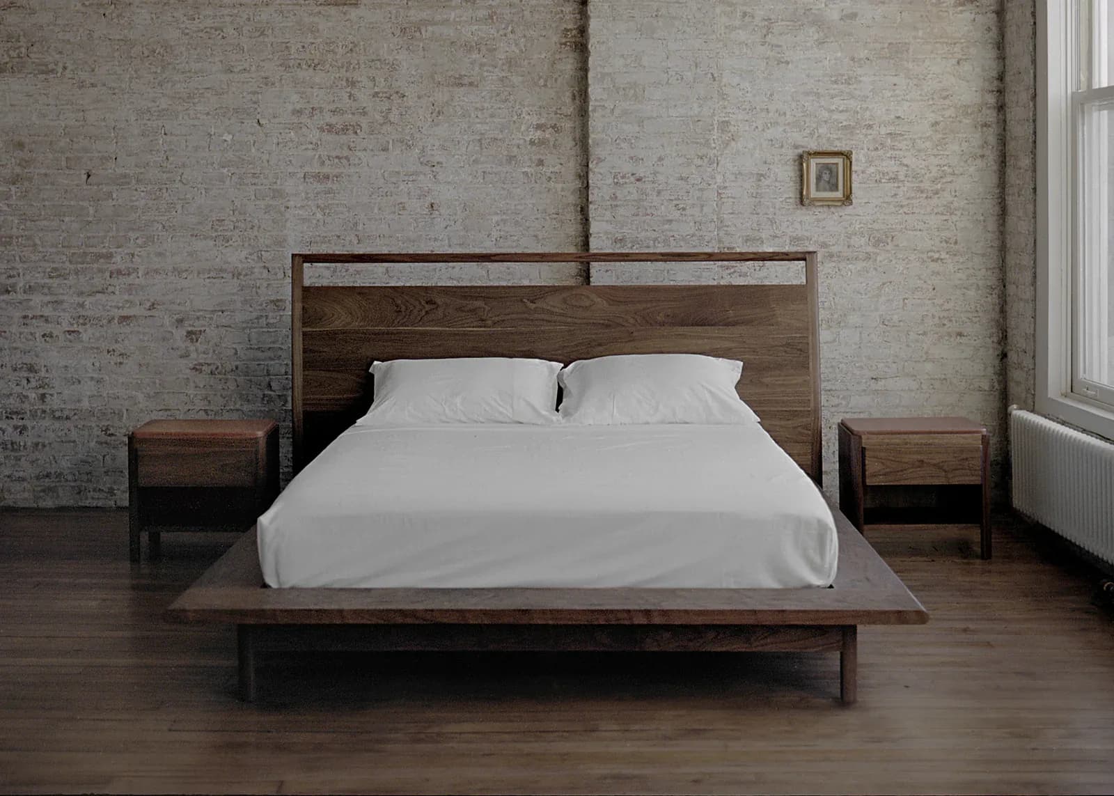 Minimalist bedroom with mid-century modern wooden bed frame, white bedding, concrete brick walls, dark wood flooring, and paired nightstands by Bowen Liu Studio.